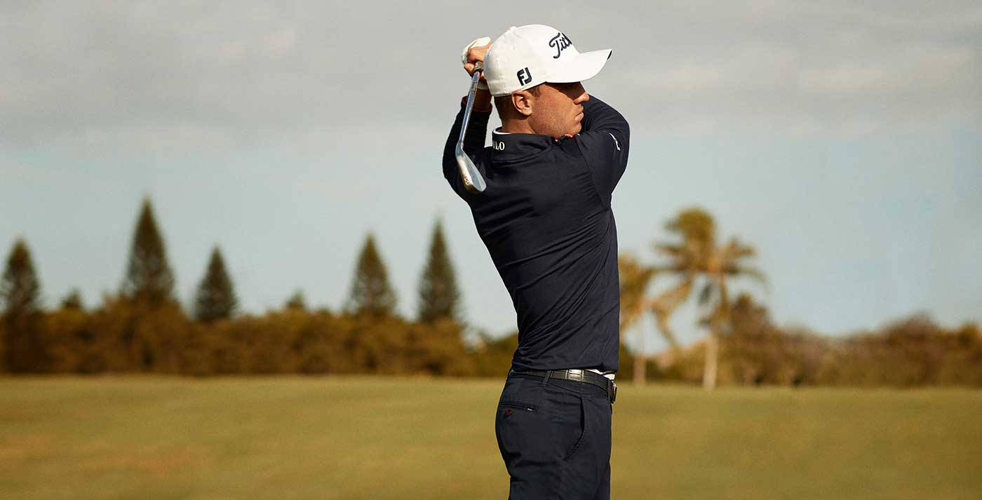Justin Thomas 高爾夫揮杆側影 壁紙 — 一位身穿海軍藍襯衫和白色帽子的高爾夫球手，在多雲的天空下，以棕櫚樹和常青樹為背景，在草地上揮桿。