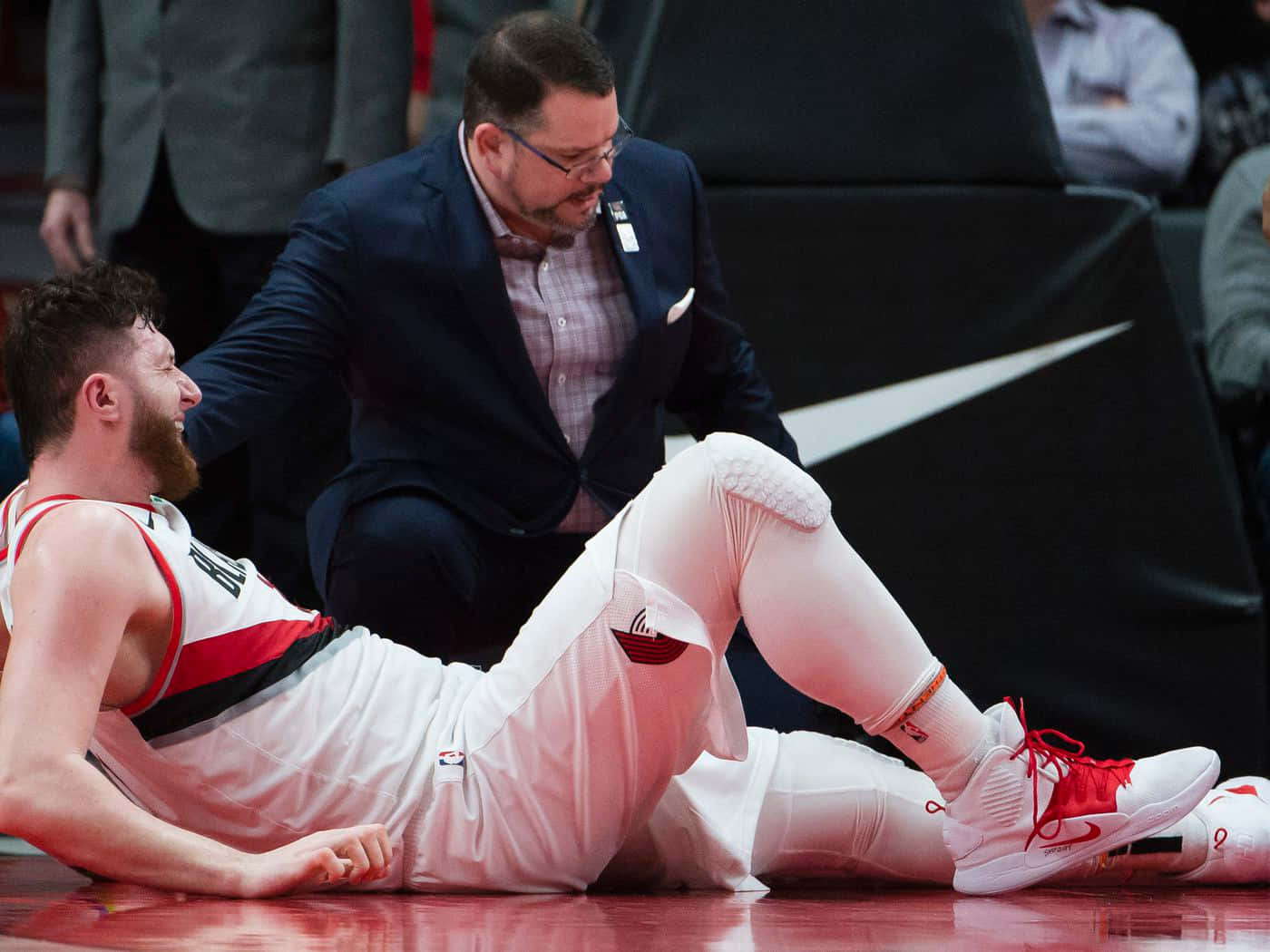 Jusuf Nurkic 帶傷奮戰 壁紙 — 一名身穿白色和紅色球衣的籃球運動員躺在球場上，一名身穿西裝的教練跪在他旁邊提供支持。