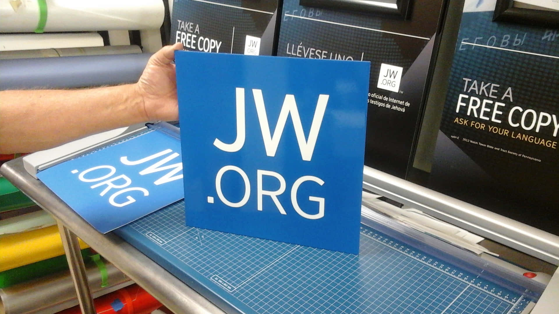 在打印店裡，一隻手拿著藍色JW.ORG標誌放在切割墊上。