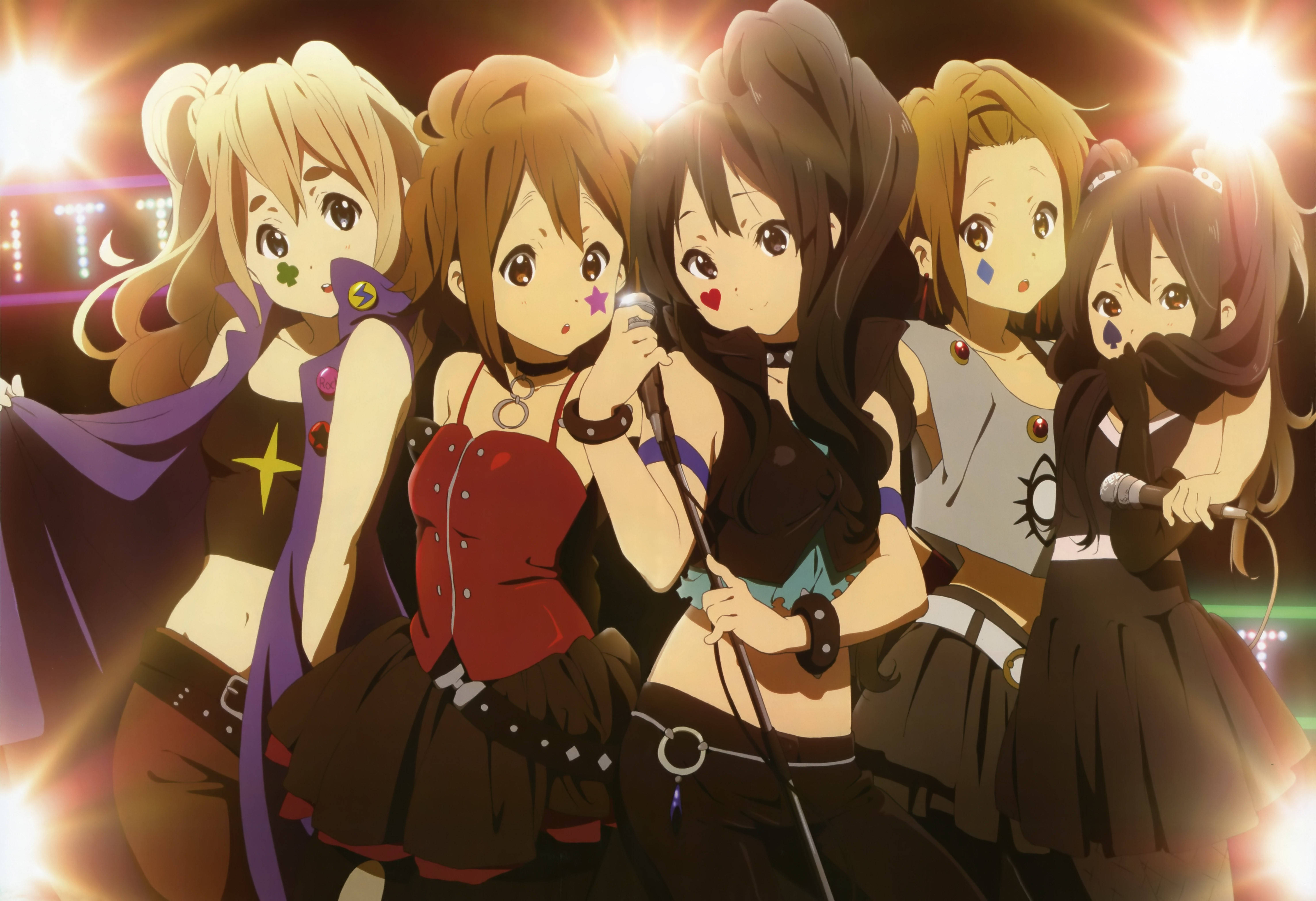 K-on 女孩穿搖滾演出服 壁紙 — 五位身穿舞台服裝的動漫女孩在明亮的聚光燈下拿著麥克風在音樂會場景中擺姿勢。