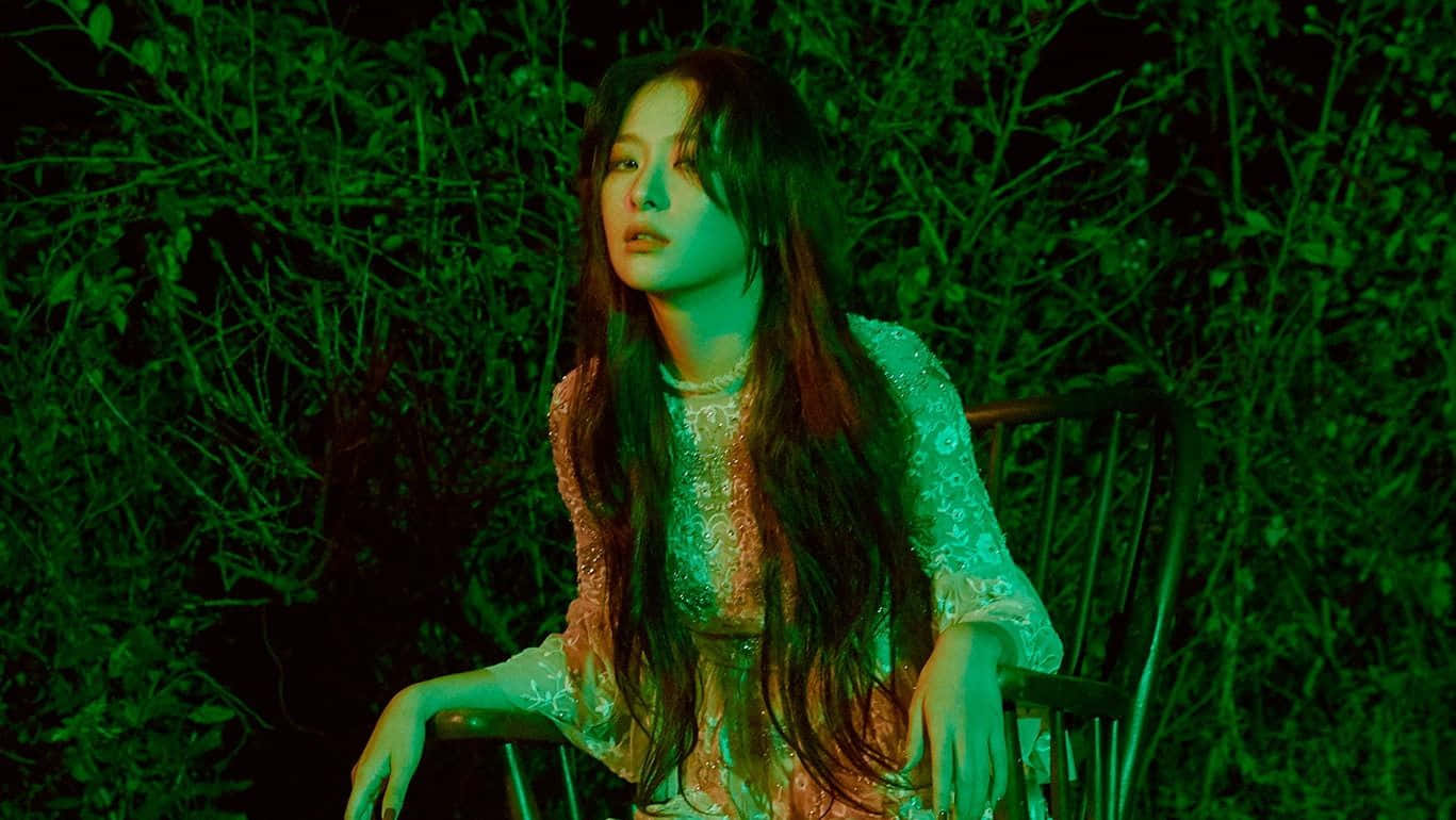 K-pop 偶像登台演出 壁紙 — 一位長黑髮的女性坐在椅子上，周圍是茂密的綠色樹葉，夜晚在詭異的綠色燈光下。