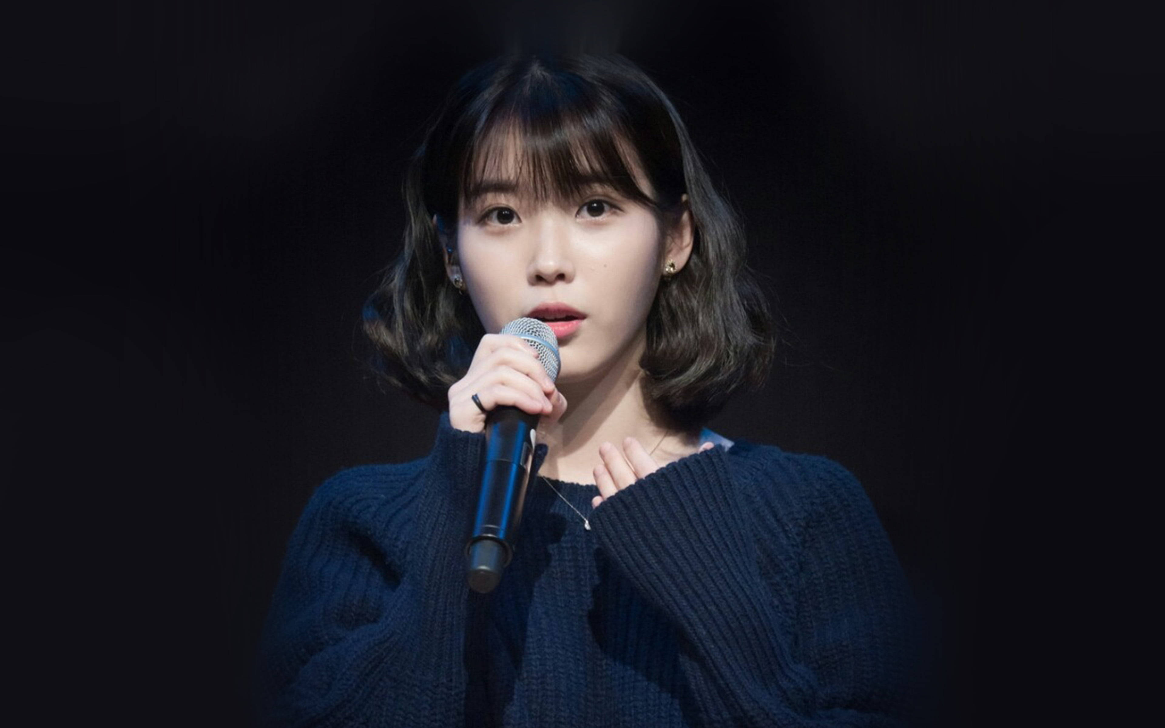 K-pop偶像李知恩 壁紙 — 一位短黑髮的年輕女性在戲劇性的舞台燈光下，穿著深藍色針織毛衣對著麥克風唱歌。