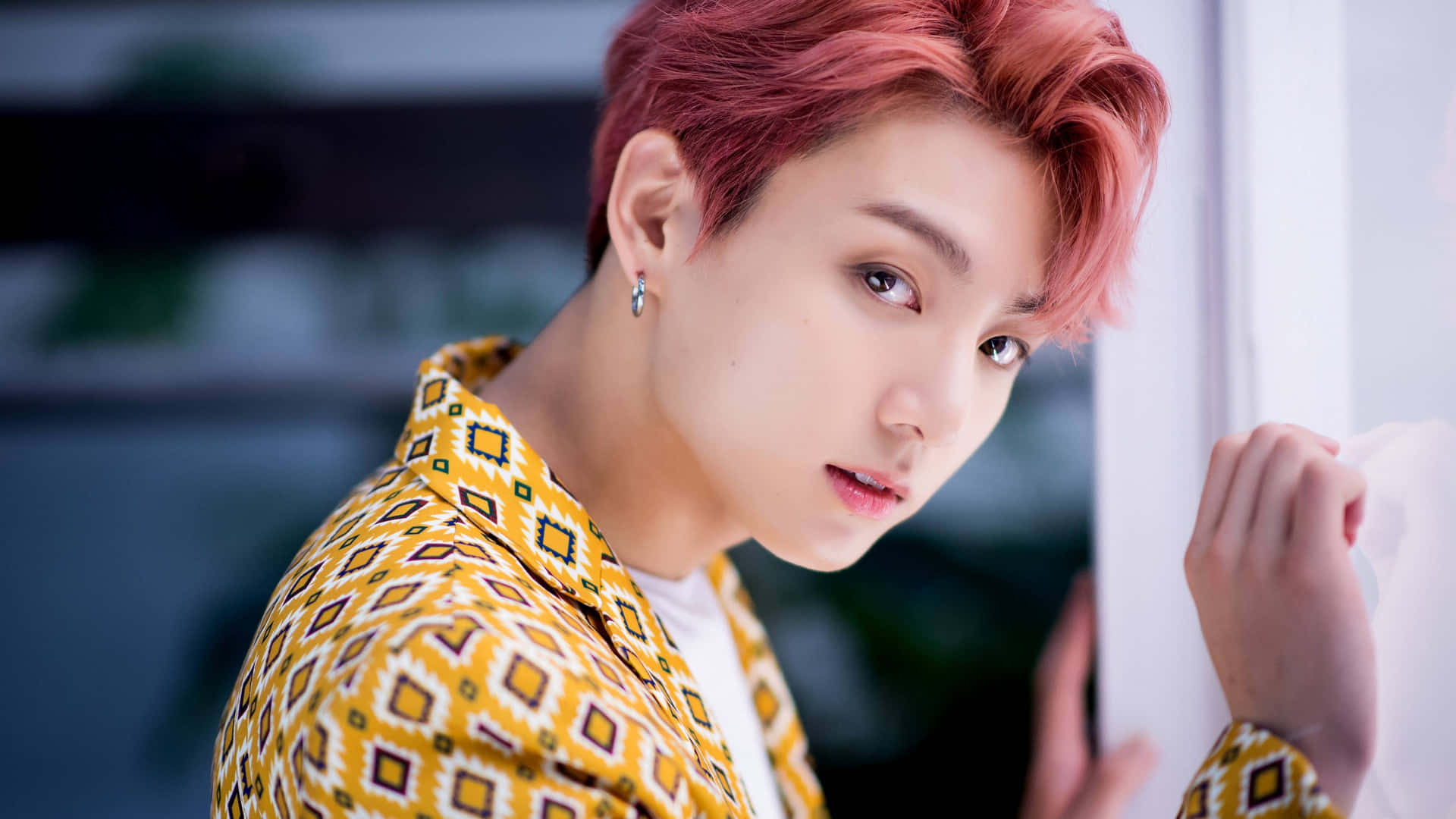 K-pop團體BTS的歌手兼詞曲創作人Jungkook拍攝寫真 壁紙 — 一位年輕男子，擁有鮮豔的粉紅色頭髮和幾何圖案的黃色襯衫，靠著白牆專注地凝視。