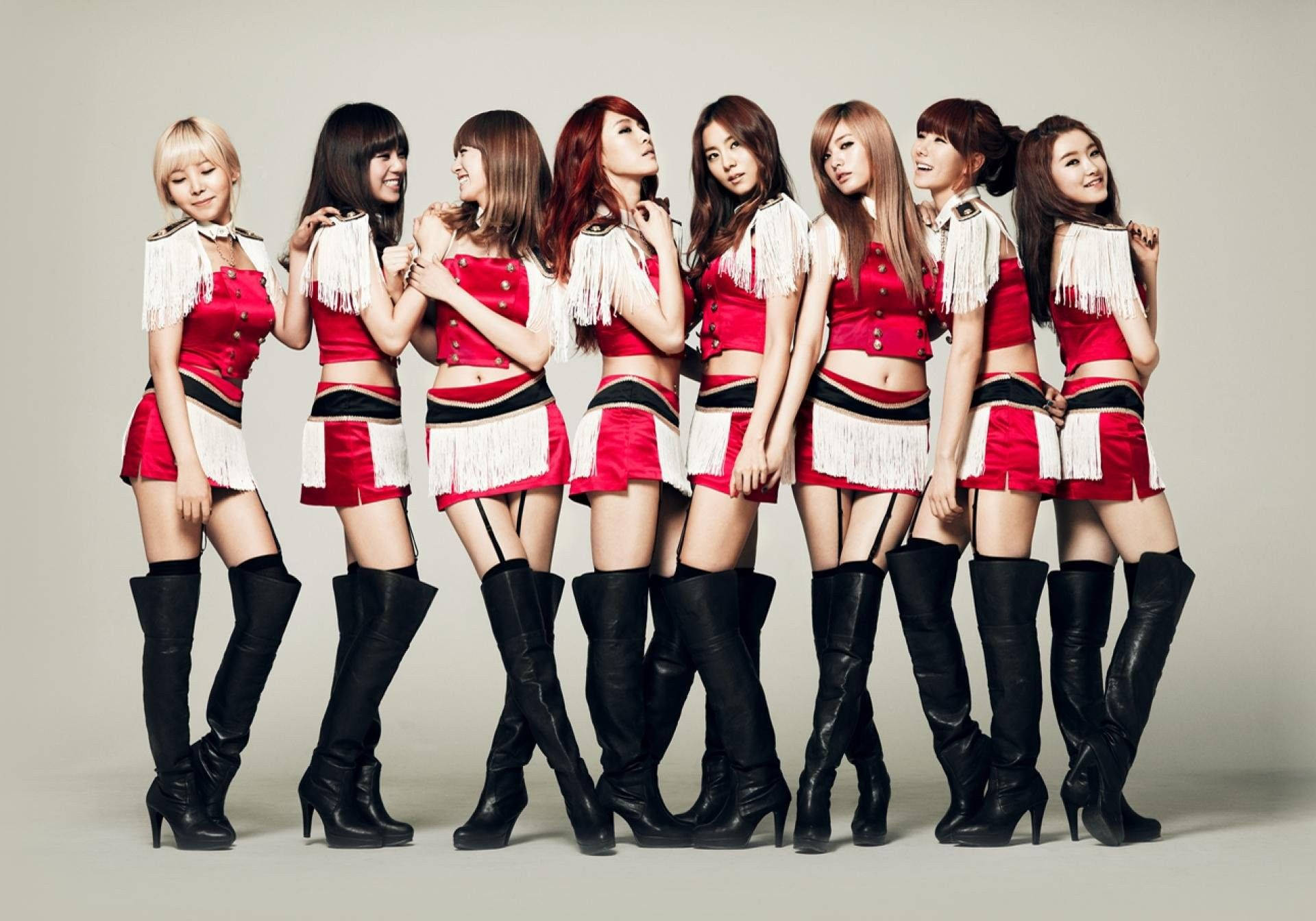 K-pop團體演唱會-聚光燈演出 壁紙 — 七位女性穿著相配的紅色流蘇服裝和黑色過膝靴，在純灰色工作室背景前自信地擺姿勢。