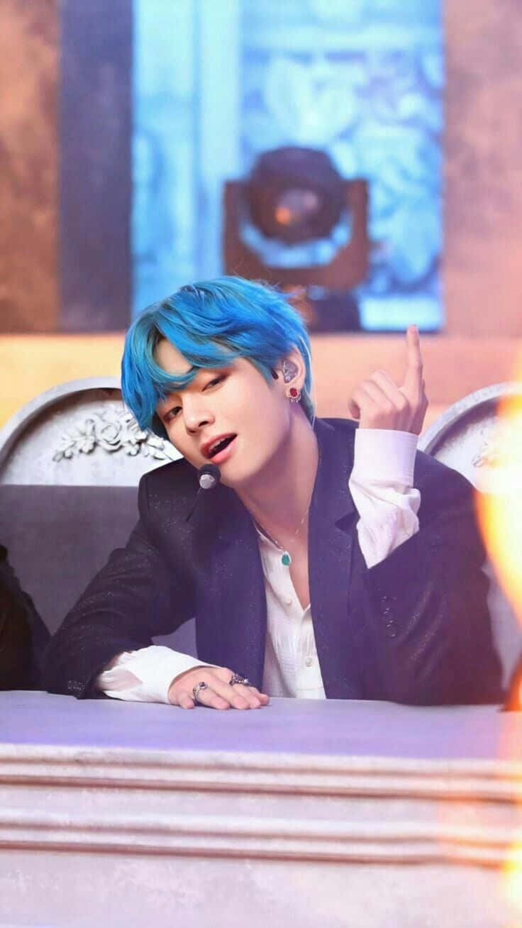 K-pop壁紙：熱門組合與偶像 壁紙 — 一位擁有鮮豔藍色頭髮的年輕男子，在溫暖的舞台燈光下坐在舞台桌前做手勢。
