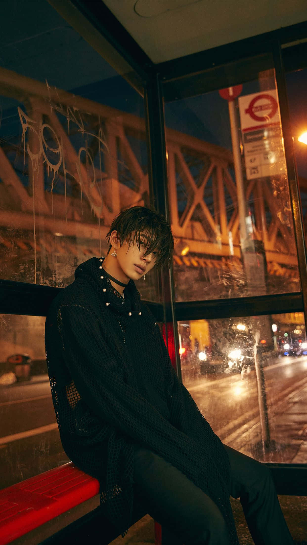 K-pop帥氣男孩照片 壁紙 — 一個穿黑色衣服的年輕人晚上坐在被雨淋濕的巴士站內的紅色長椅上，外面有塗鴉和城市燈光閃爍。