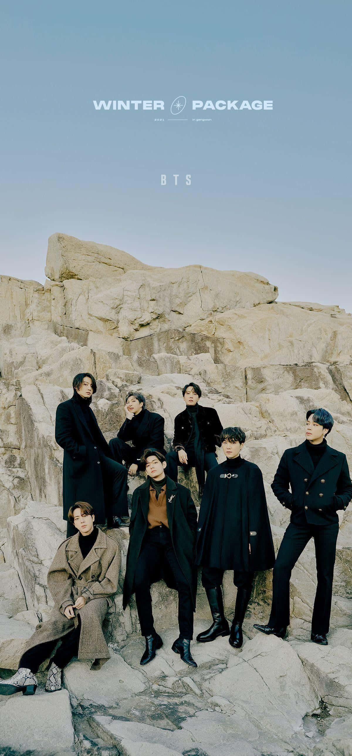 K-pop組合登台演唱熱門歌曲 壁紙 — 七位男士身穿時尚深色大衣，在淡藍色天空下，自信地站在崎嶇的岩石懸崖上。