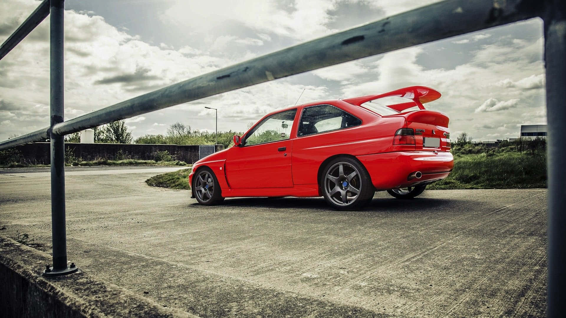 Ford Escort 桌布 壁紙 — 一架鮮紅色嘅跑車，帶有大型尾翼，停泊喺金屬欄杆框住嘅陰天之下。