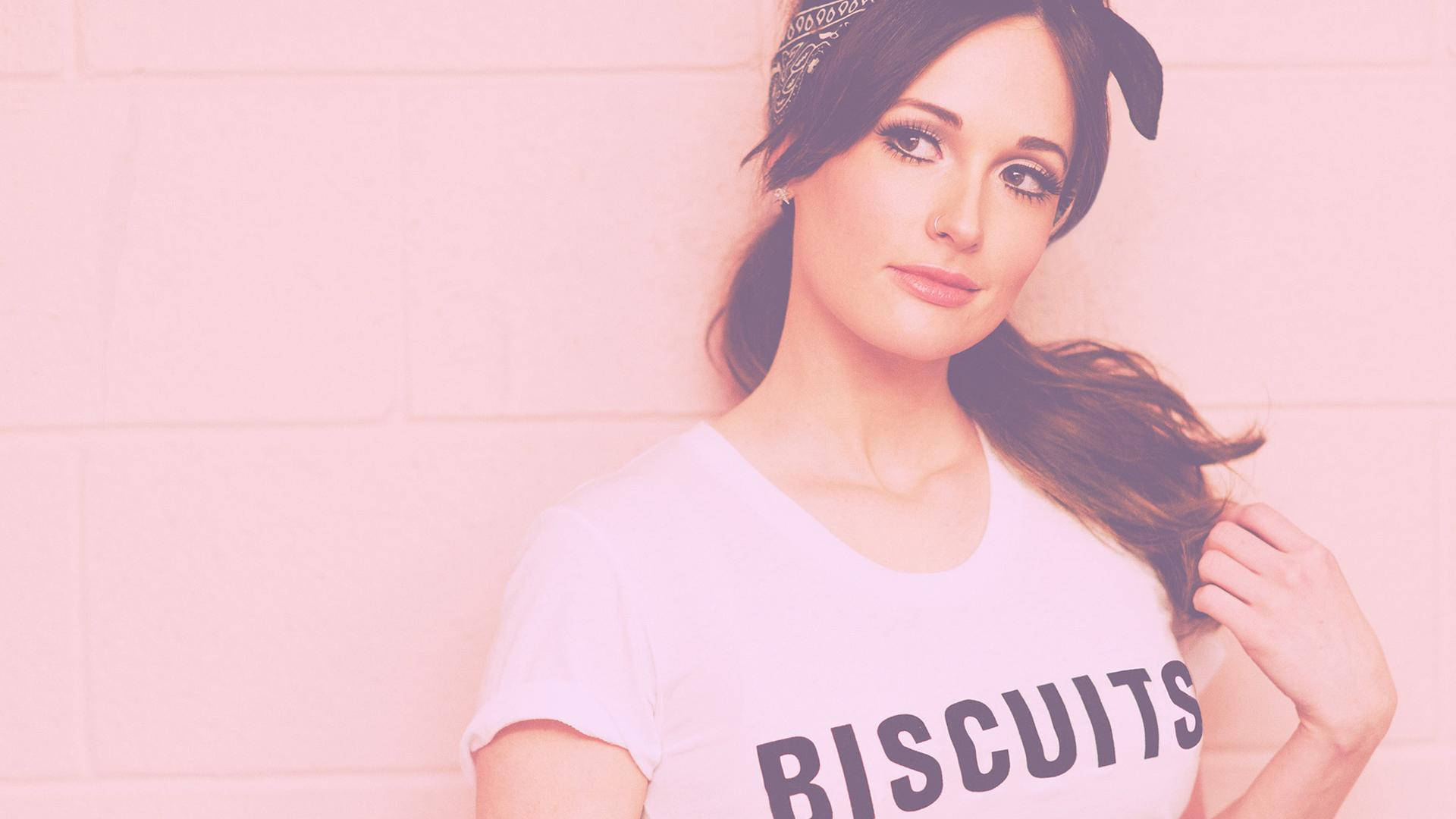 Kacey Musgraves 淡粉少女風 壁紙 — 一位深色頭髮、戴頭巾的女性，身穿印有biscuits字樣的白色T恤，靠著粉紅磚牆。