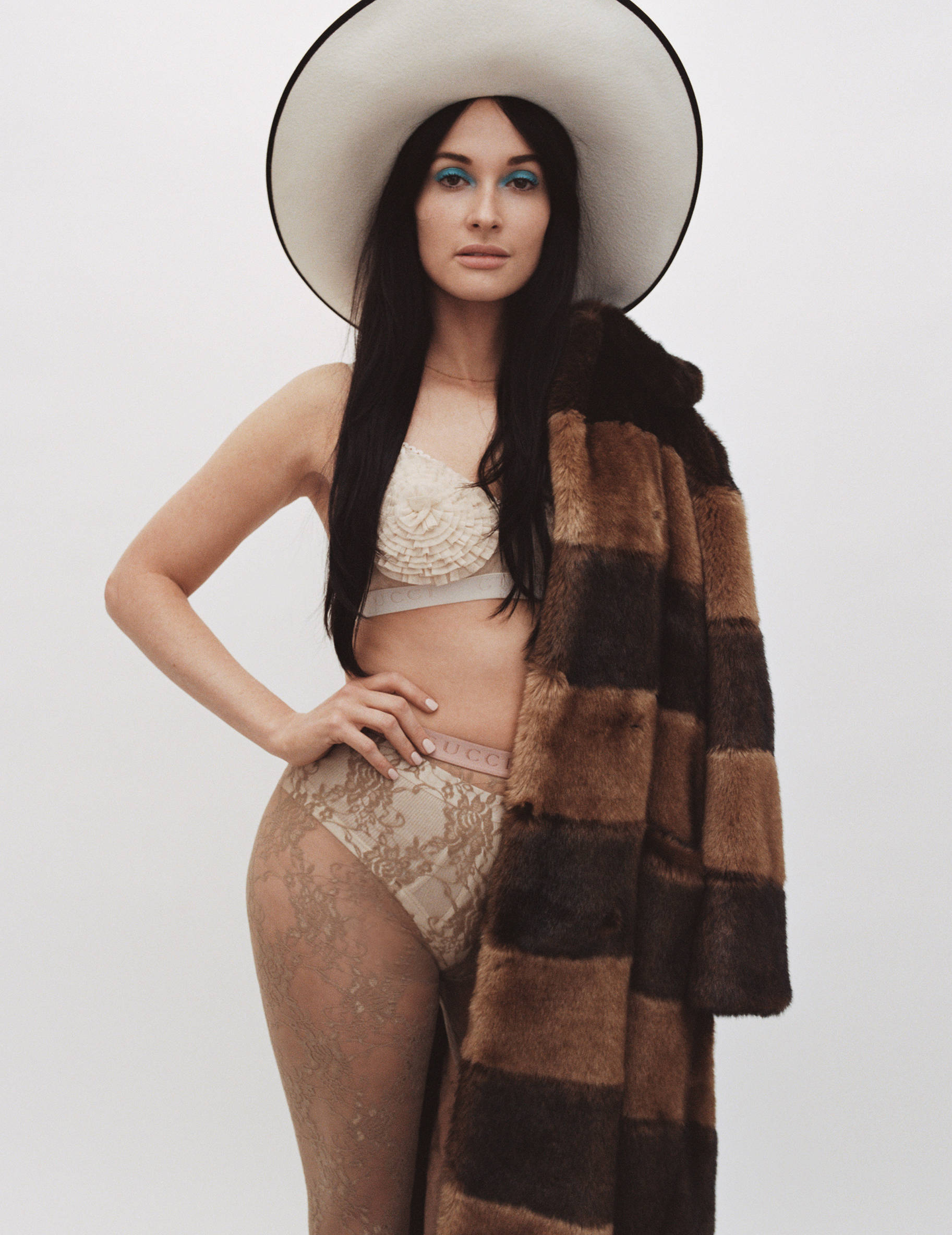 Kacey Musgraves Vogue 預告拍攝 壁紙 — 一位戴著寬邊帽和皮草大衣的女性，在純白背景下自信地穿著內衣擺姿勢。