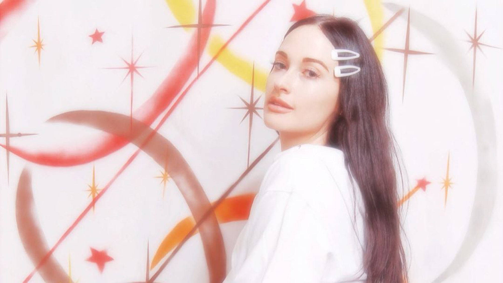 Kacey Musgraves 夢幻拍攝 壁紙 — 一位擁有長長黑髮和白色夾子的女性，在充滿活力的紅、橙、黃色星星和漩渦的抽象背景前溫柔地凝視。