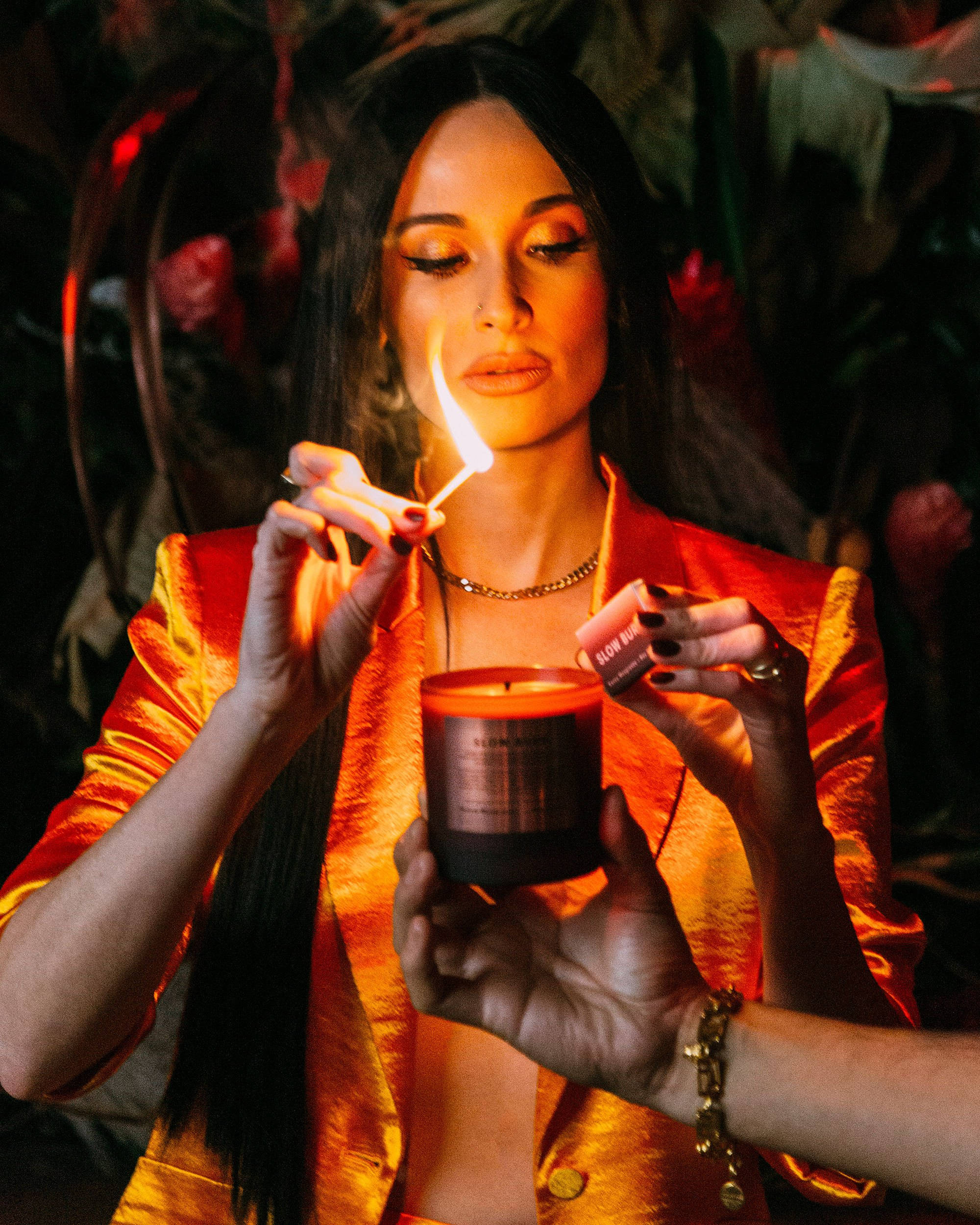 Kacey Musgraves 燭光點燃 壁紙 — 一位身穿閃亮橙色外套的女性在光線昏暗、佈滿鮮花的房間裡用火柴點燃蠟燭。