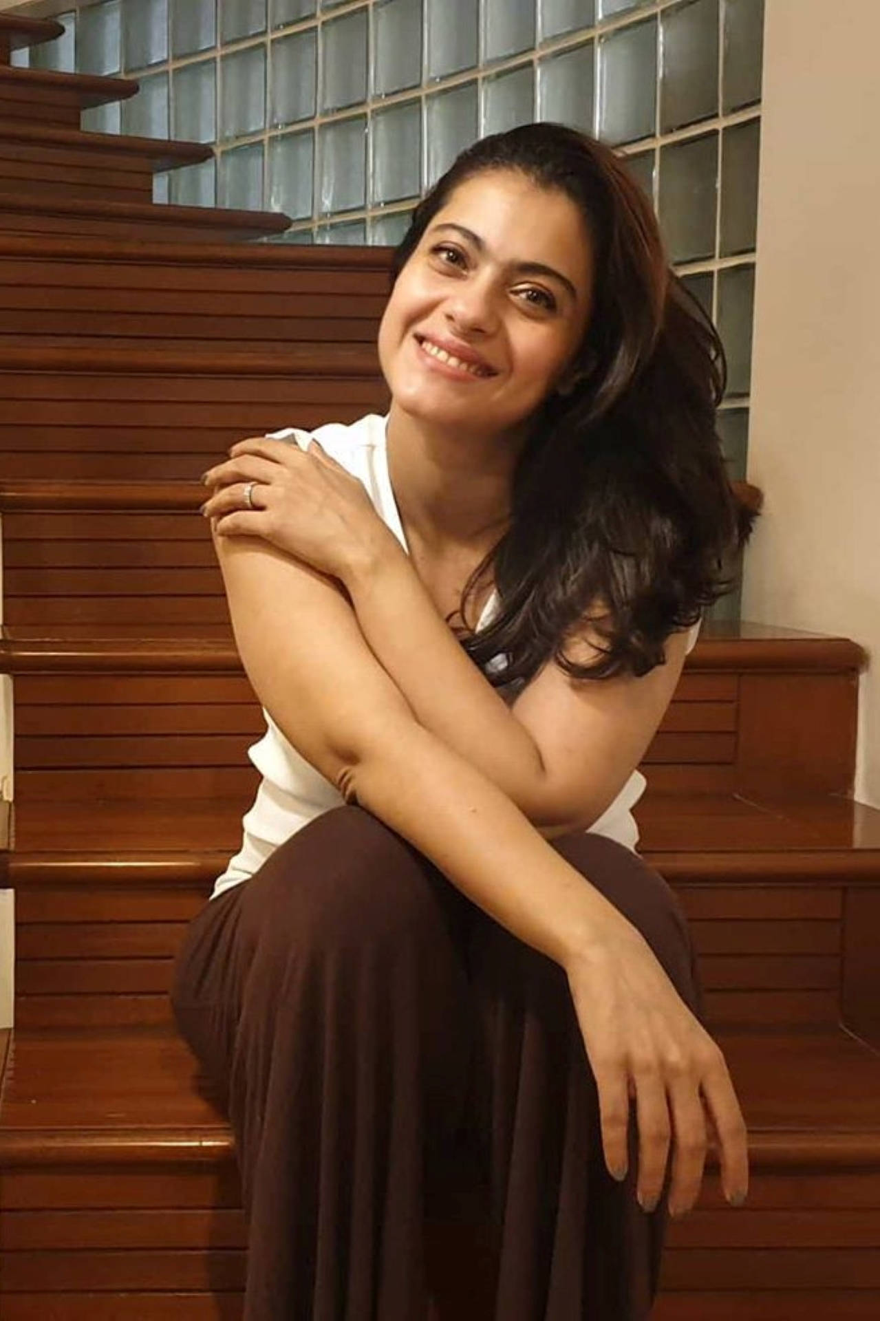 Kajol 自然美肌照 壁紙 — 一位深色波浪髮的女性在玻璃磚牆前的木樓梯上盤腿而坐，露出溫暖的笑容。