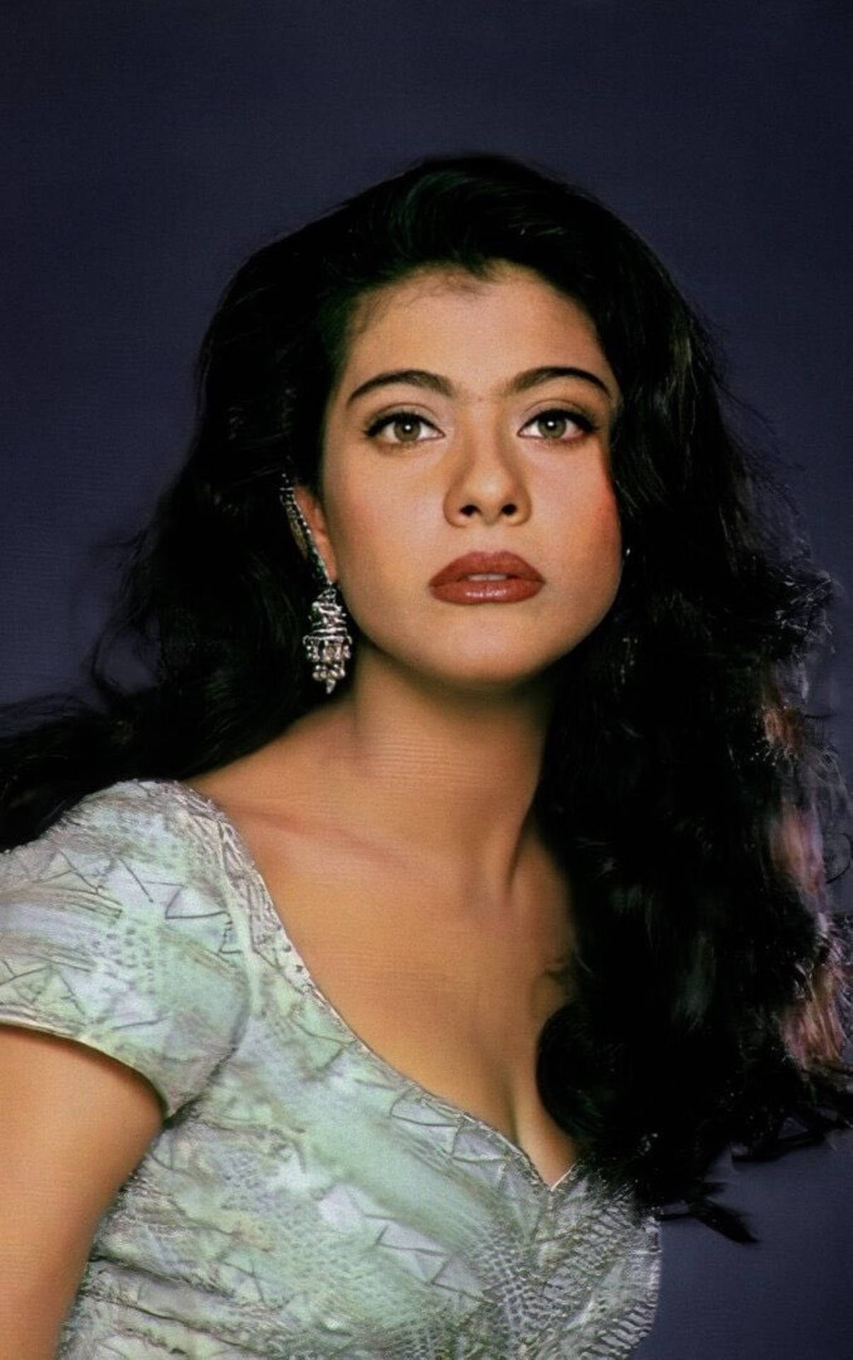 Kajol 身著傳統服飾的優雅風采 壁紙 — 一位擁有深色波浪長髮和華麗耳環的女性，穿著淺綠色圖案上衣，在深紫色背景下凝視前方。