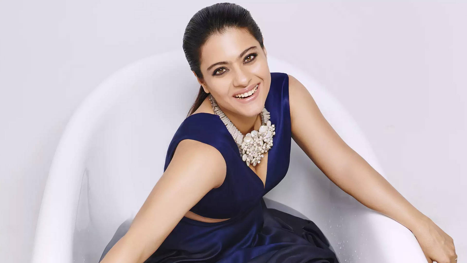 Kajol 身著深藍時尚造型 壁紙 — 一位身穿皇家藍色緞面連衣裙的容光煥發的女性，坐在時尚的白色椅子上，對著柔和的灰色背景溫暖地微笑。