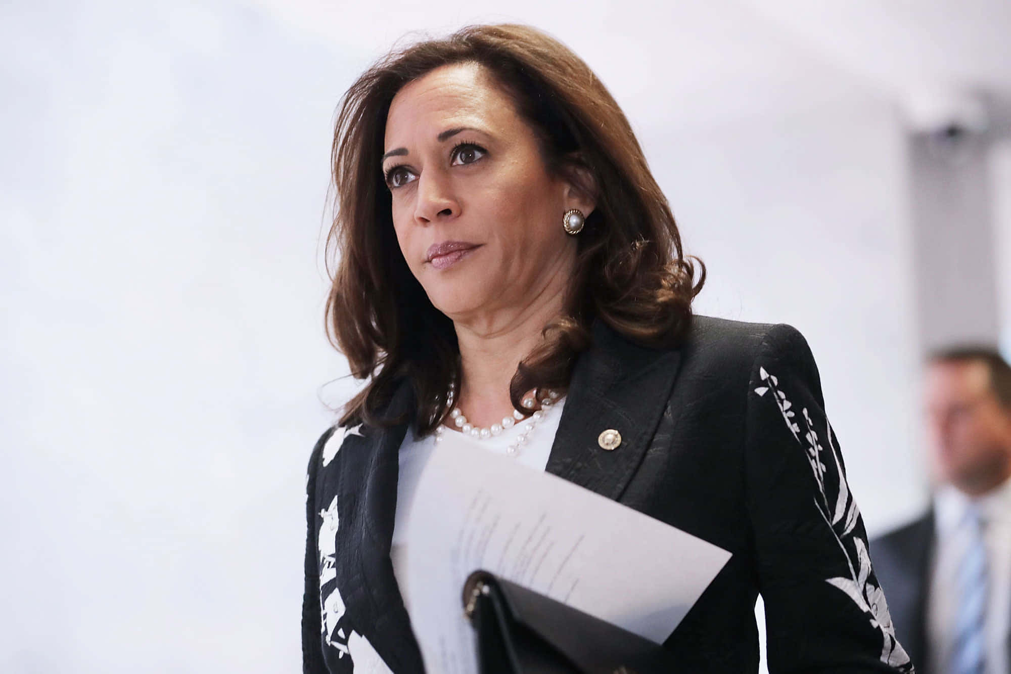 Kamala Harris 桌布 壁紙 — Kamala Harris 身穿黑色花卉西裝外套，在明亮的室內環境中手持文件，表情嚴肅。