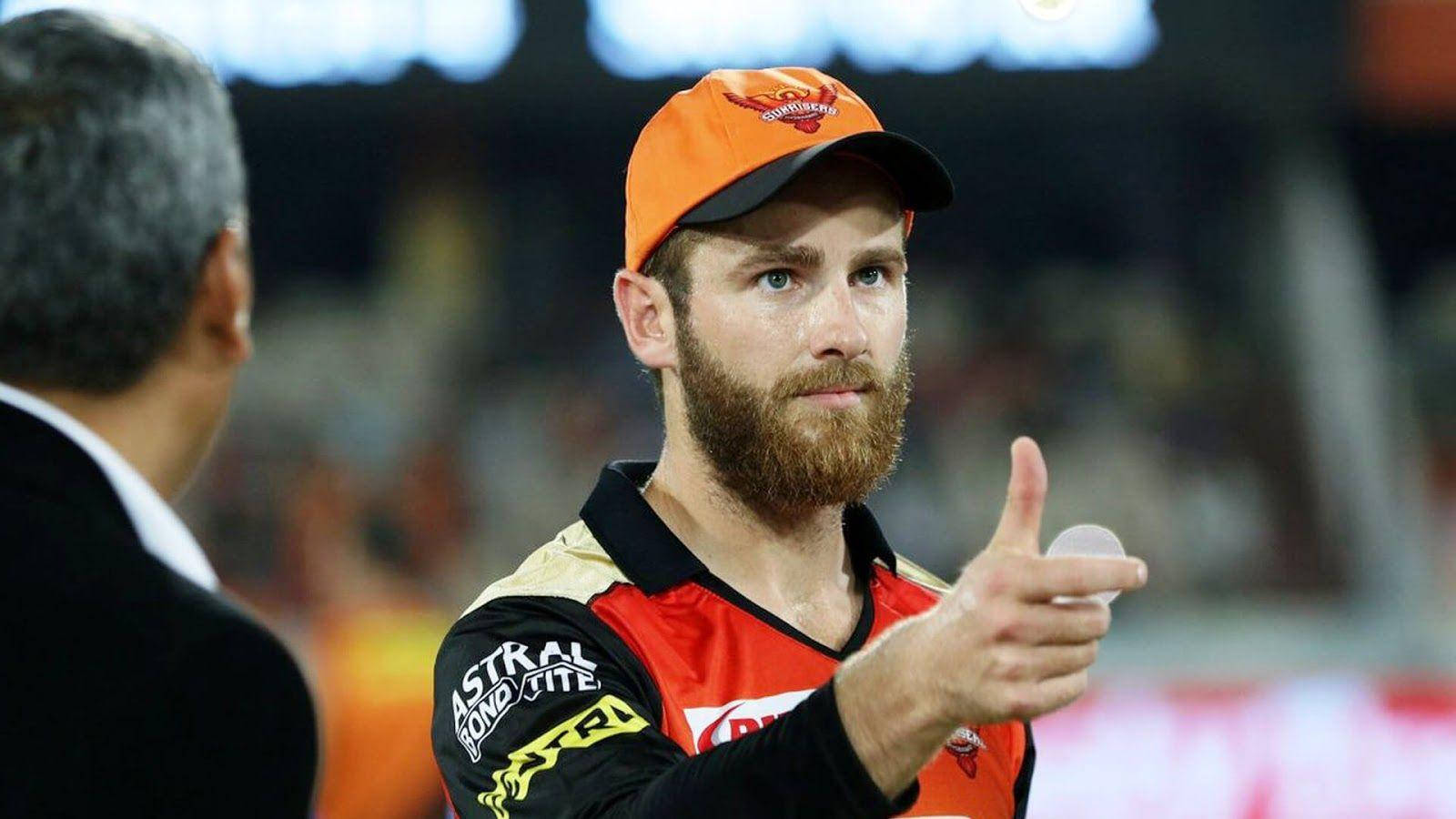 Kane Williamson 橙色帽子 壁紙 — 一位留鬍鬚的板球運動員，戴著橙色帽子和球衣，在夜間比賽中在體育場燈光下豎起大拇指。