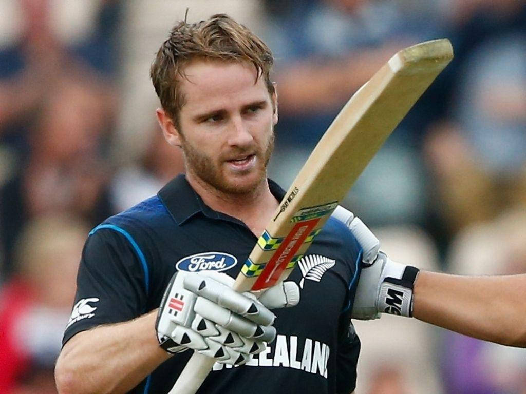 Kane Williamson 拿板球棒 壁紙 — 一位專注的新西蘭板球運動員在比賽中緊握球棒，面對模糊的體育場人群，展現出強烈的決心。