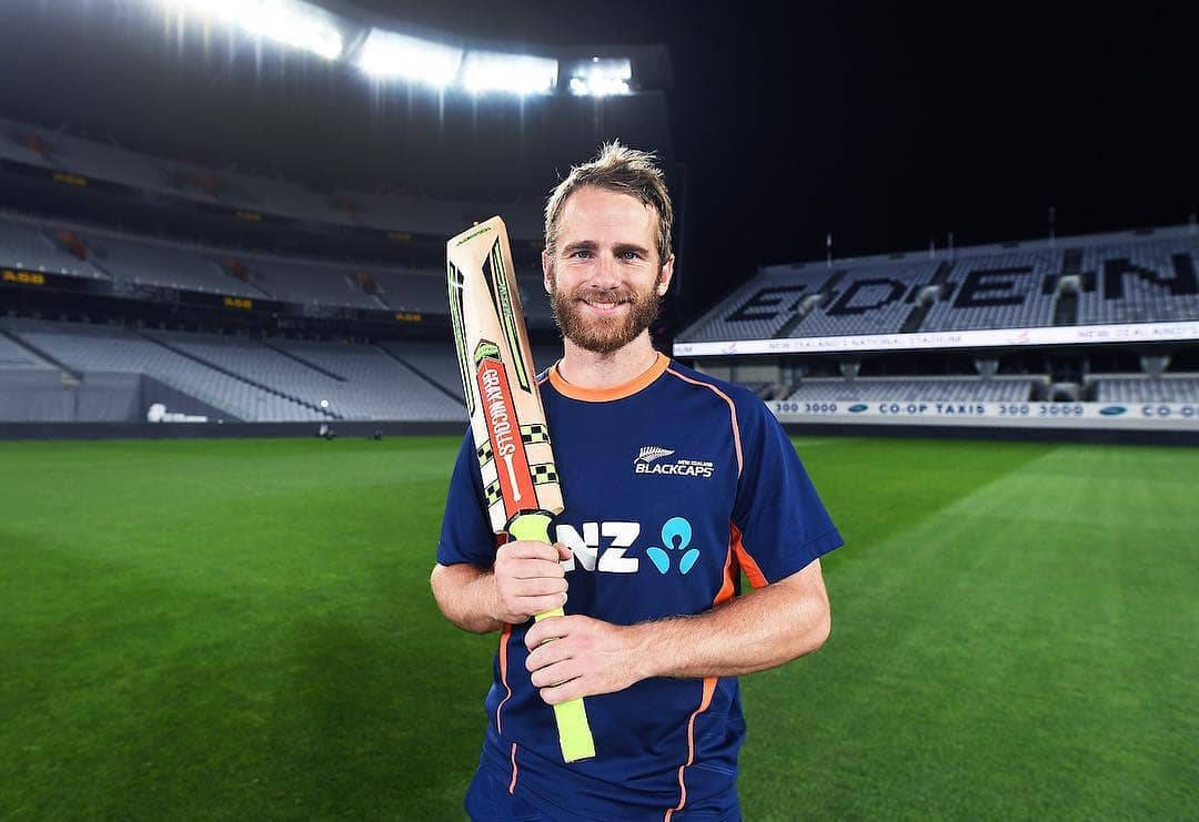 Kane Williamson 板球場夜間揮棒 壁紙 — 一位微笑留鬍鬚的板球運動員身穿新西蘭黑帽隊球衣，在夜間燈光明亮的球場上手持板球拍。