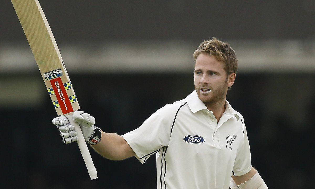 Kane Williamson 拿板球棒 壁紙 — 一名身穿白色球衣的板球手高舉球棒，臉上帶著勝利的表情，背景是模糊的體育場。