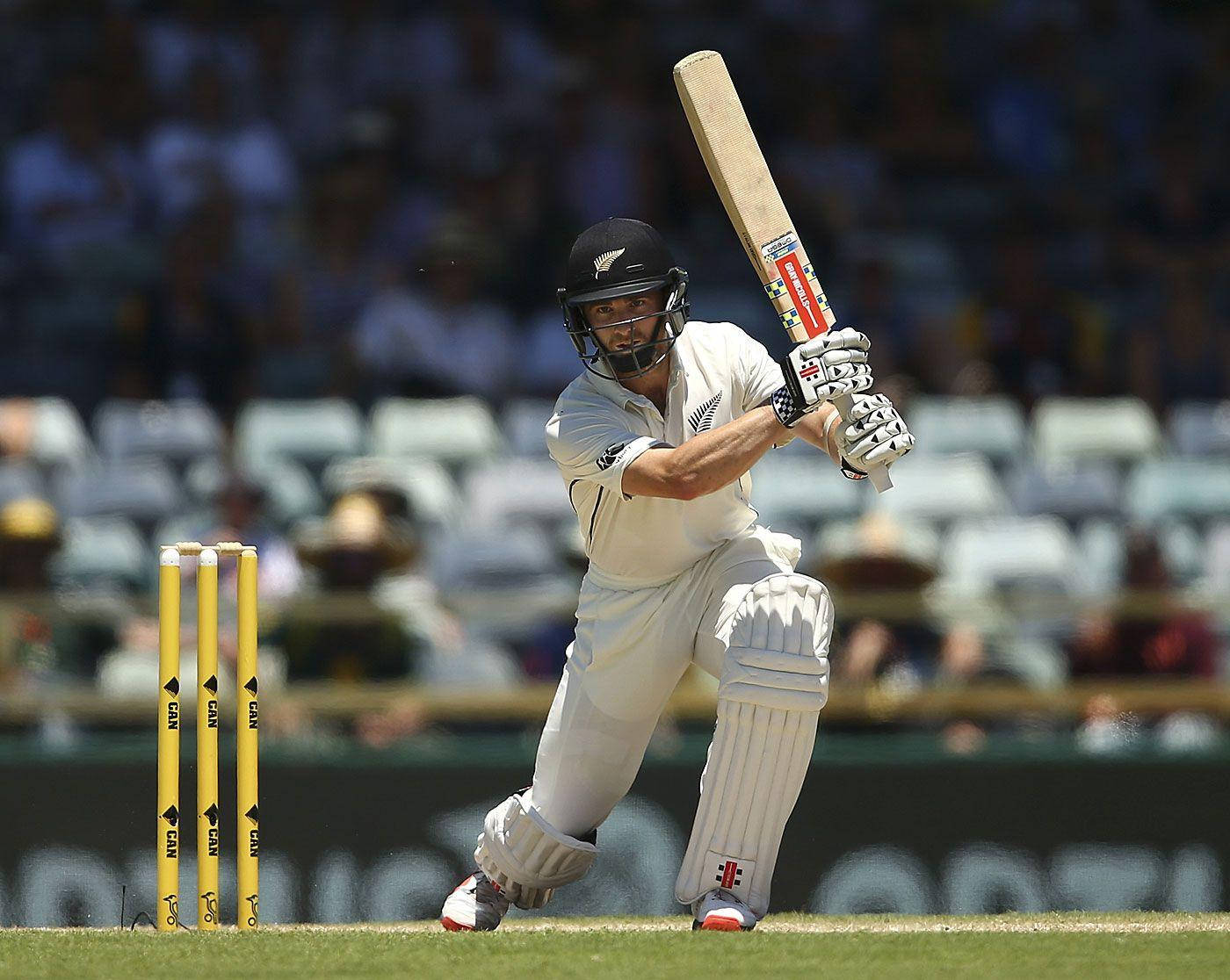 Kane Williamson 板球員 壁紙 — 一名身穿白色球衣的板球擊球手在陽光明媚的綠色球場上，強力揮動球棒擊向靠近黃色三柱門的球。
