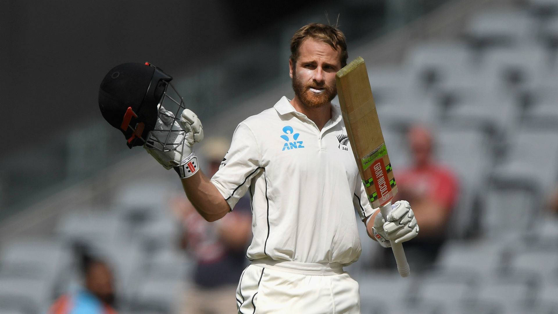 Kane Williamson 頭盔與球棒 壁紙 — 一名穿著白色制服的板球運動員在明亮的日光下走在體育場上，手持球棒和頭盔。