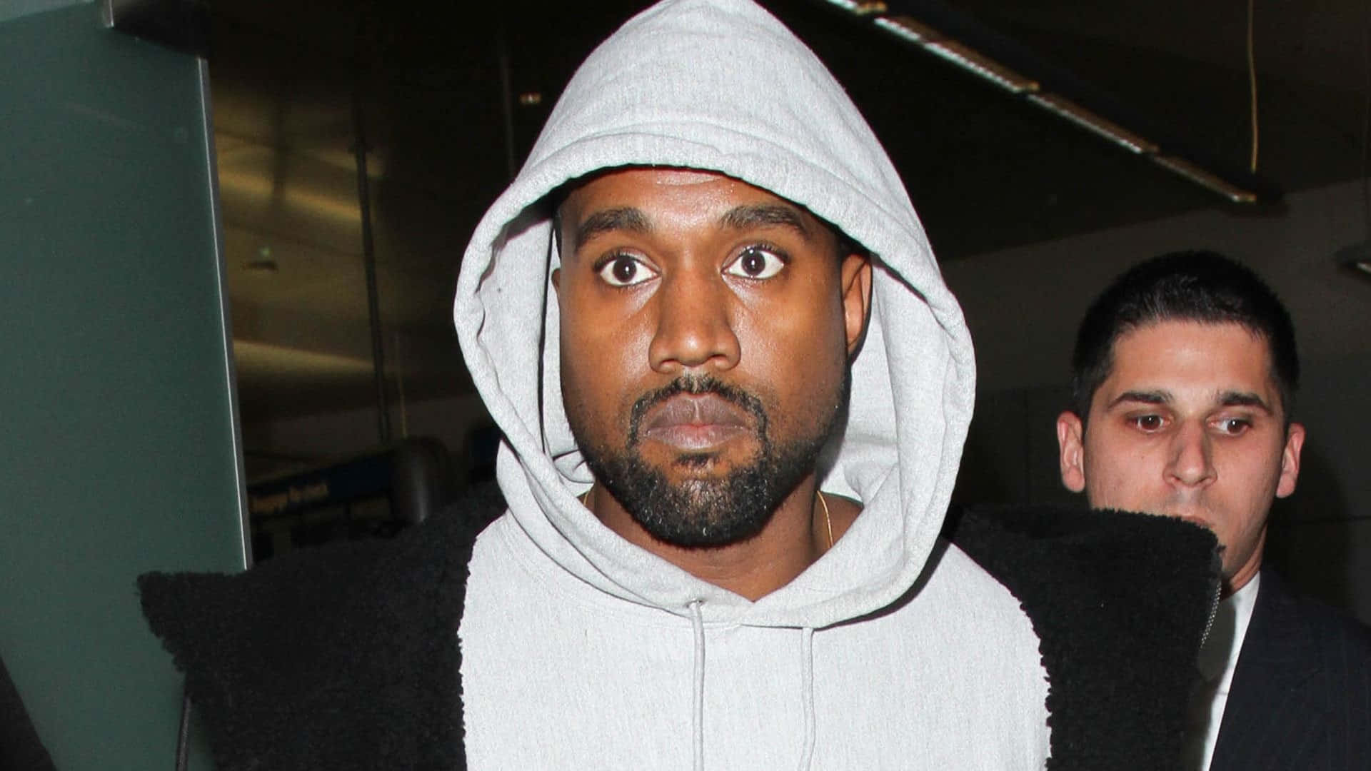  壁紙 — Kanye West 身穿灰色連帽衫，表情驚訝，站在光線昏暗的室內環境中。