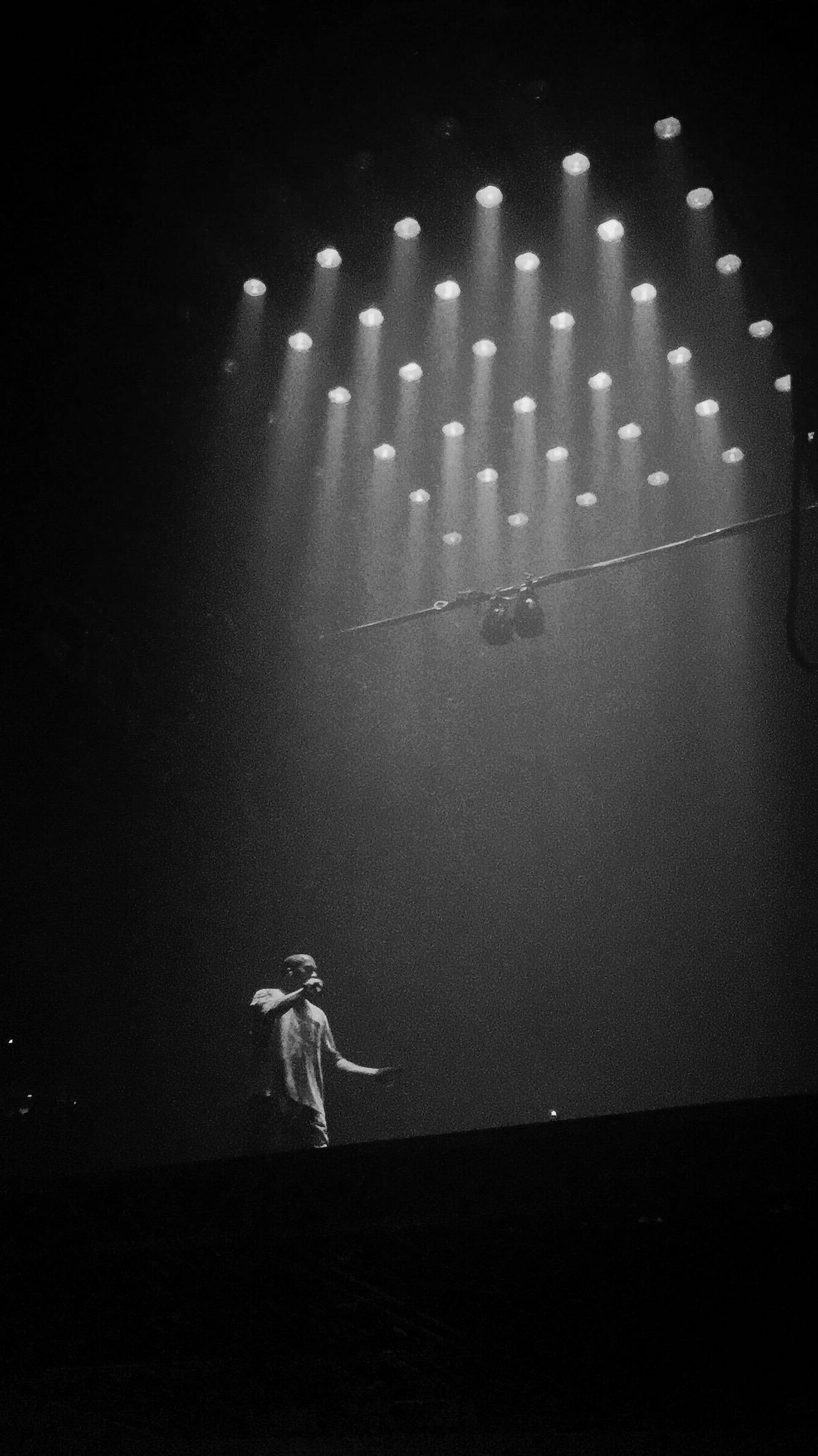Kanye West聚光燈下演出 壁紙 — 一位孤獨的表演者站在燈光昏暗的舞台上，沐浴著從上方投射而來的戲劇性聚光燈。