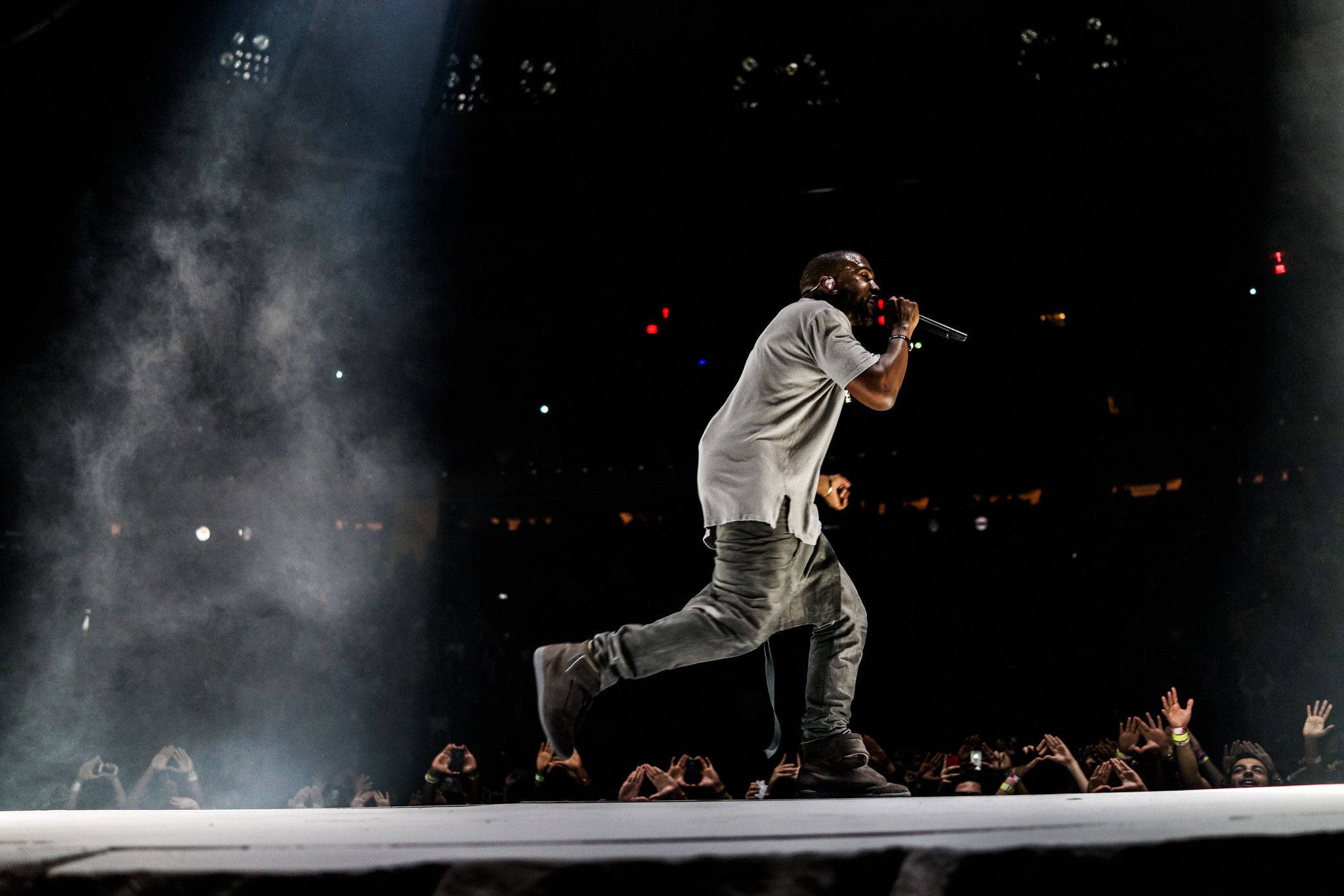 Kanye West舞台踱步演唱 壁紙 — 表演者在煙霧彌漫的舞台上，在明亮的聚光燈下動態地大步走，下方有投入的觀眾。