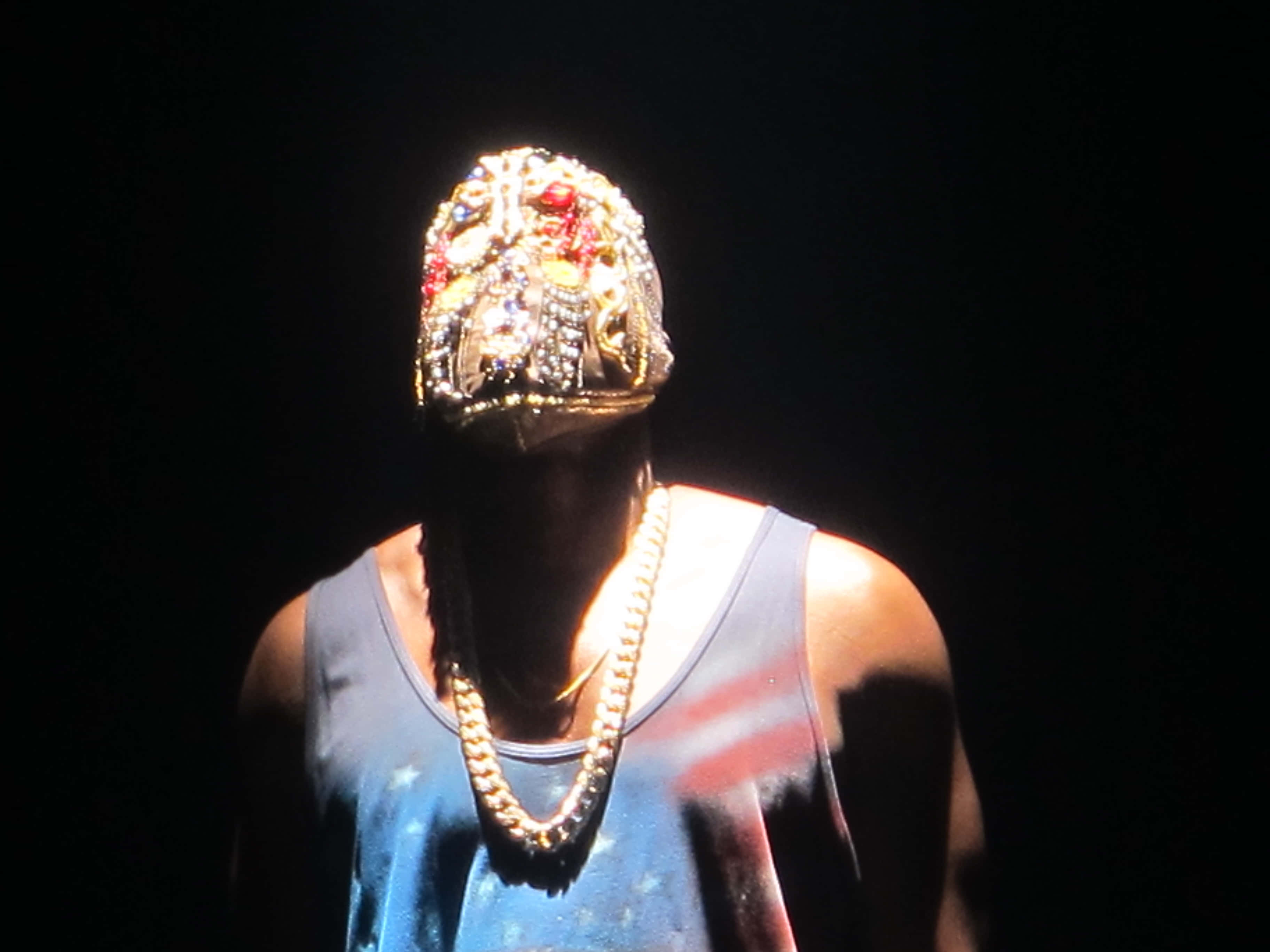 Kanye West 登台 Yeezus 巡迴演出 壁紙 — 一個人戴著鑲有寶石的面具和粗鏈條，在黑色背景下的戲劇性聚光燈下。