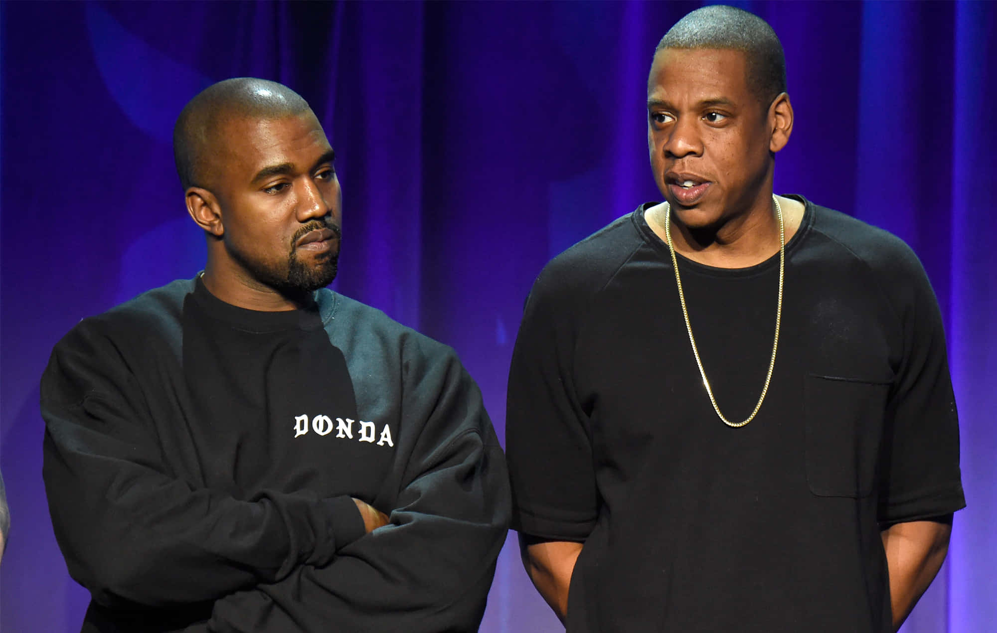 Kanye 桌布 壁紙 — Kanye West 和 Jay Z 站在舞台上，背景是紫色窗簾，一人穿著 'DONDA' 衛衣，另一人戴著金鏈。