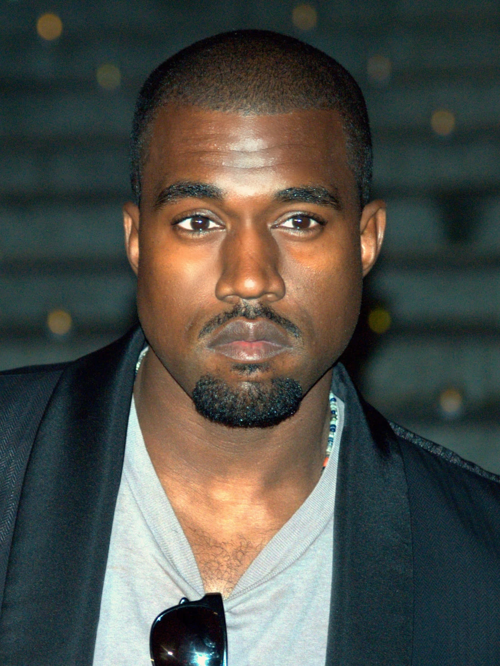 Kanye 桌布 壁紙 — Kanye West 嘅特寫肖像，表情嚴肅，身穿黑色西裝外套配灰色恤衫，太陽眼鏡掛喺領口。
