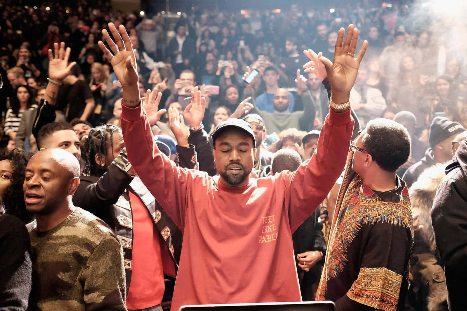 Kanye 桌布 壁紙 — Kanye West 在紅色運動衫中舉起雙手，在明亮的舞台燈光下，在歡呼的人群中舉行音樂會。