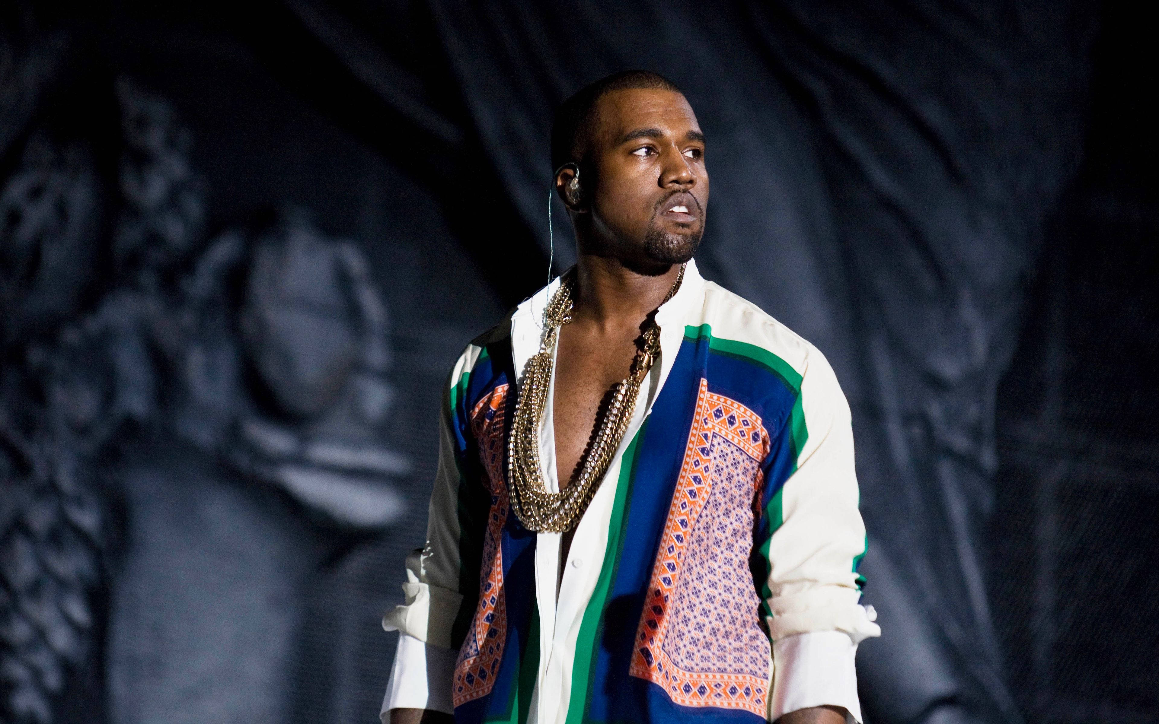 Kanye West Colourful Long Sleeves 壁紙 — Kanye West 在舞台上表演，穿著色彩繽紛的圖案外套和層疊項鍊，在戲劇性的舞台燈光下。