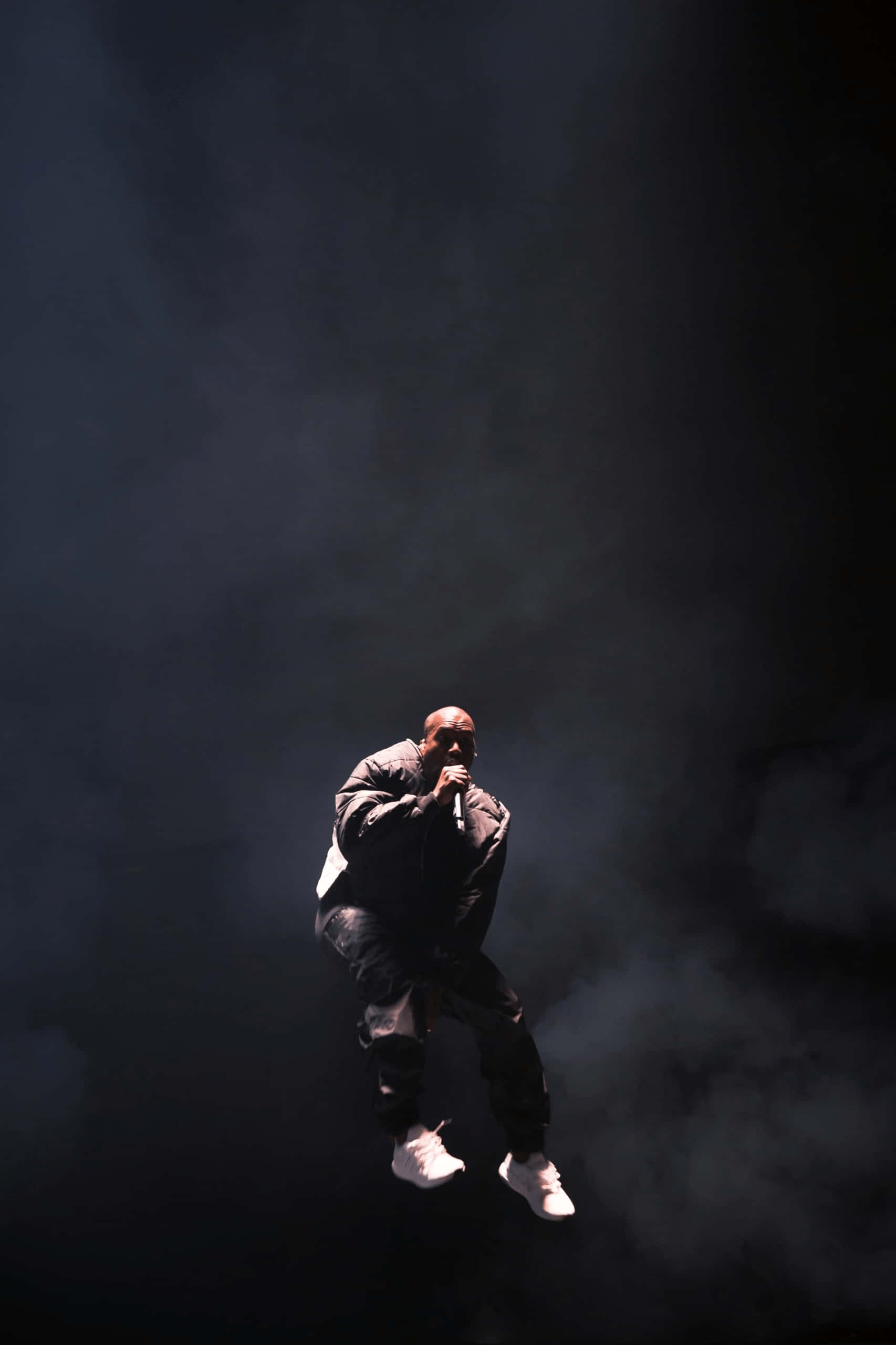Kanye West 推出全新時尚 iPhone 系列 壁紙 — 表演者在黑暗煙霧中懸浮，在戲劇性舞台燈光下手持麥克風。