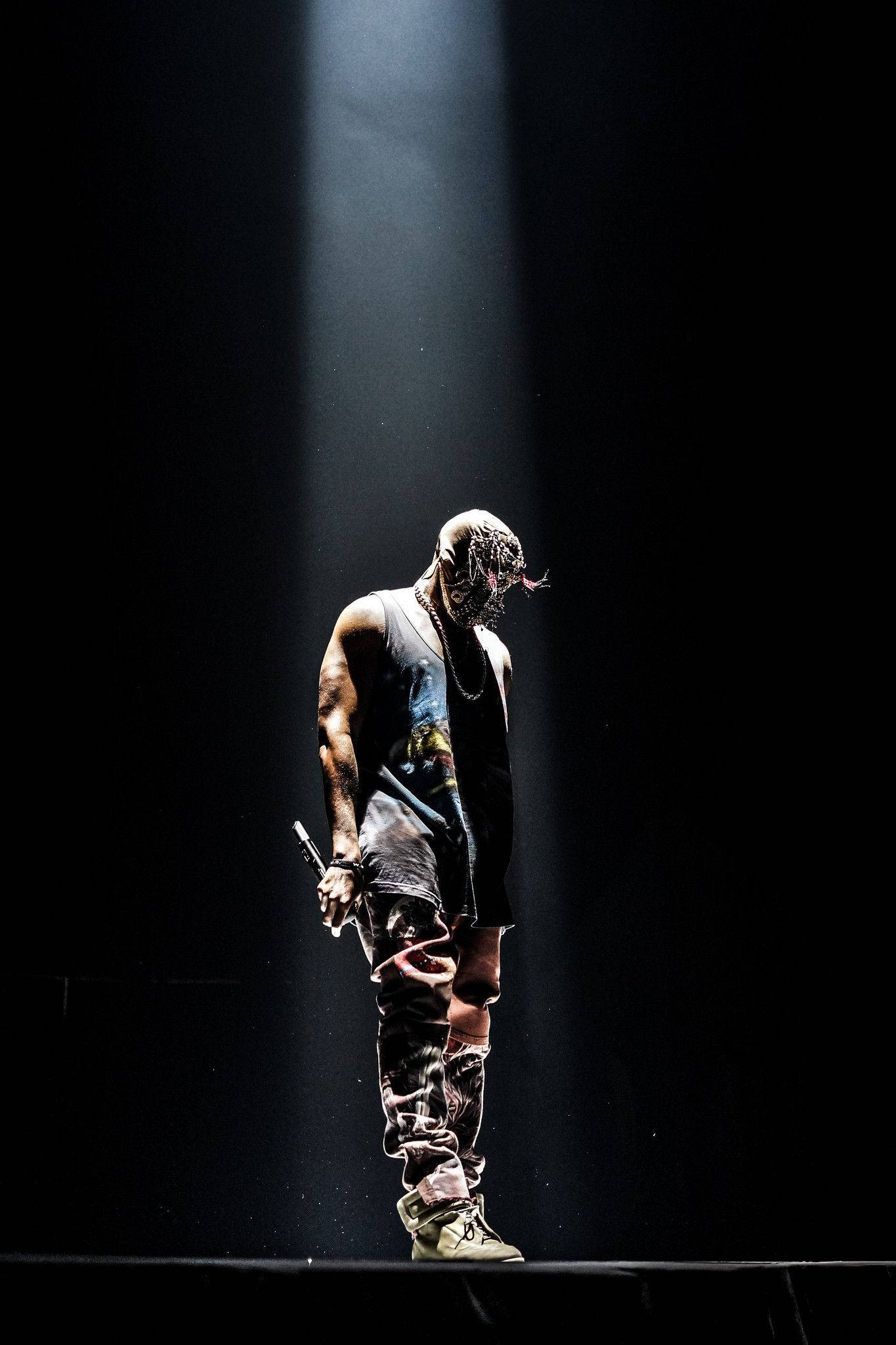 Kanye West 演唱會舞台 壁紙 — 一位戴著面具、穿著破爛服裝的表演者獨自站在黑暗舞台上的單一聚光燈下。