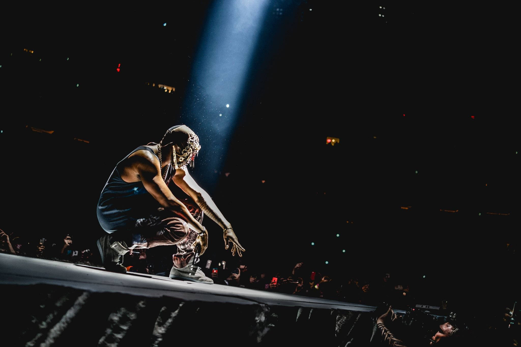 Kanye West 登台演出 壁紙 — 表演者在聚光燈下蹲在舞台上，粉絲們在黑暗的音樂會場地舉起手機。