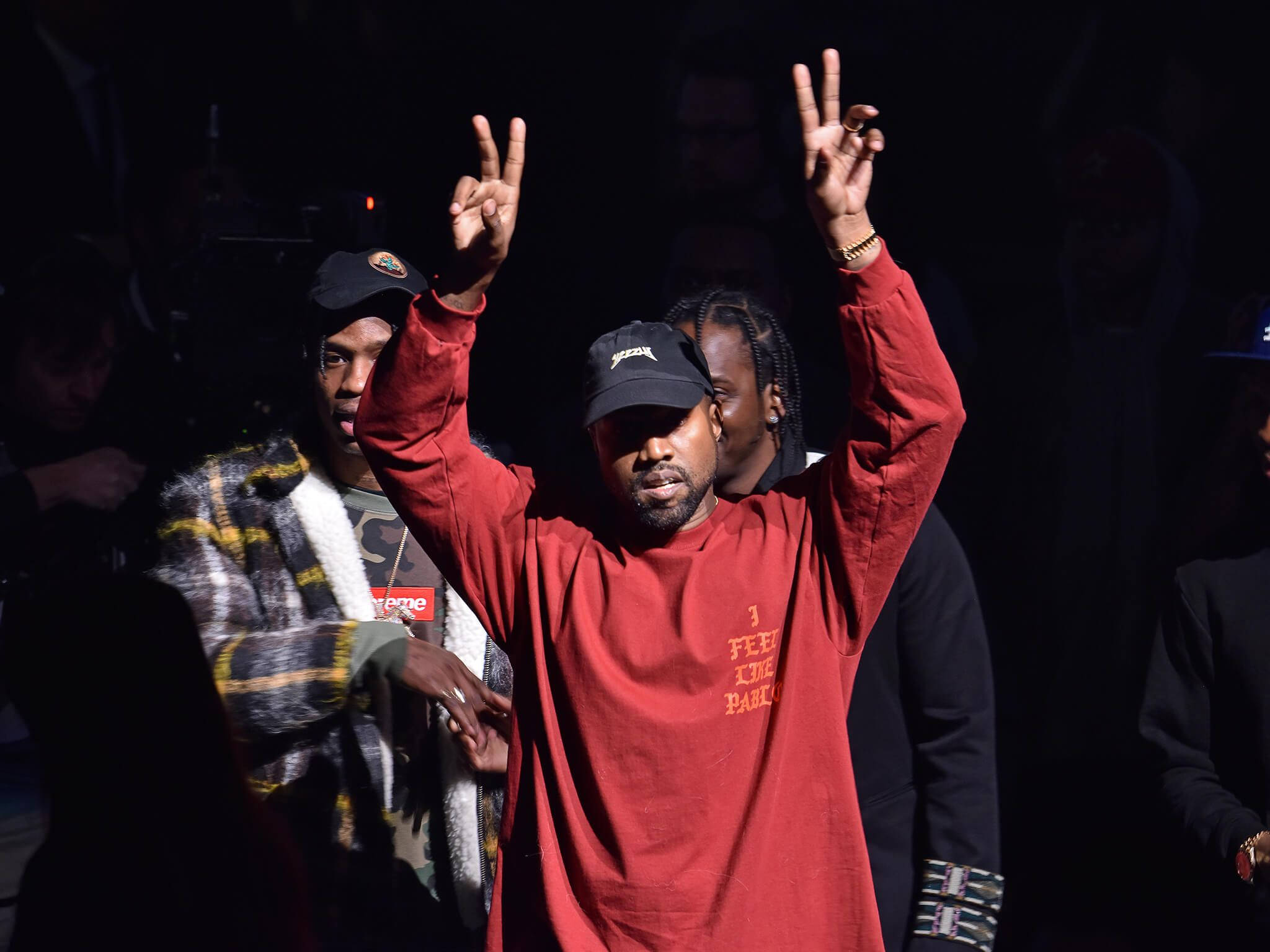 Kanye West Yeezy Peace 壁紙 — Kanye West 穿著紅色衛衣和黑色帽子，在舞台燈光下舉起雙手做出和平手勢，身後是模糊的觀眾。