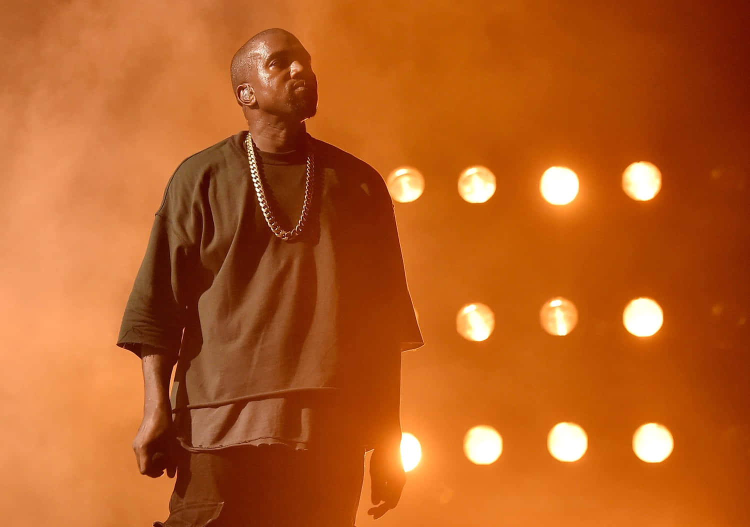 Kanye 桌布 壁紙 — Kanye West 站在舞台上，沐浴在溫暖的橙色燈光中，身後有發光的聚光燈。