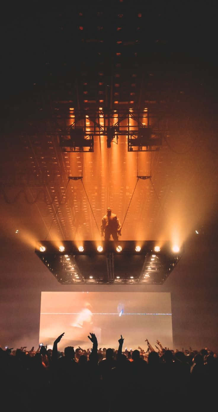 Kanye West 與他的經典 iPhone 壁紙 — 一位表演者站在一個懸掛的舞台上，沐浴在金色的燈光中，下方是擁擠的音樂廳中舉起的手海。