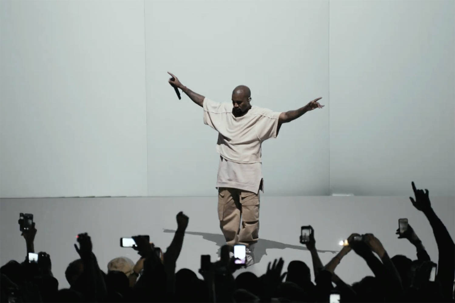 Kanye West 與母親 Donda 共享溫馨時刻 壁紙 — 一位身穿米色服裝的表演者站在舞台中央，雙臂伸展，而下面的觀眾舉起手機捕捉這一刻。
