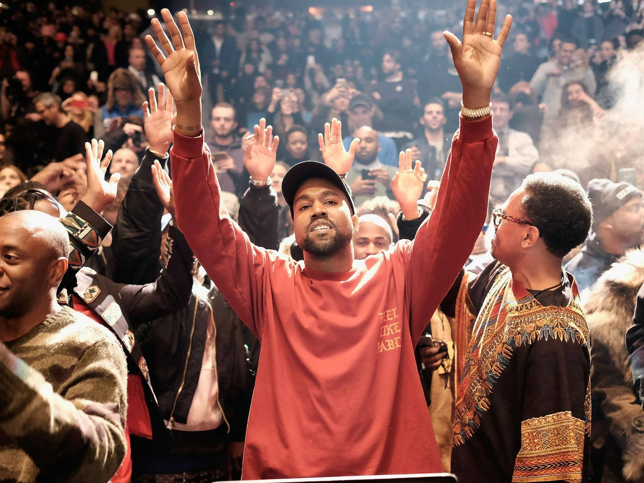 Kanye West 與觀眾群 壁紙 — Kanye West 在演唱會中於煙霧和閃爍燈光下在人群中高舉雙手。