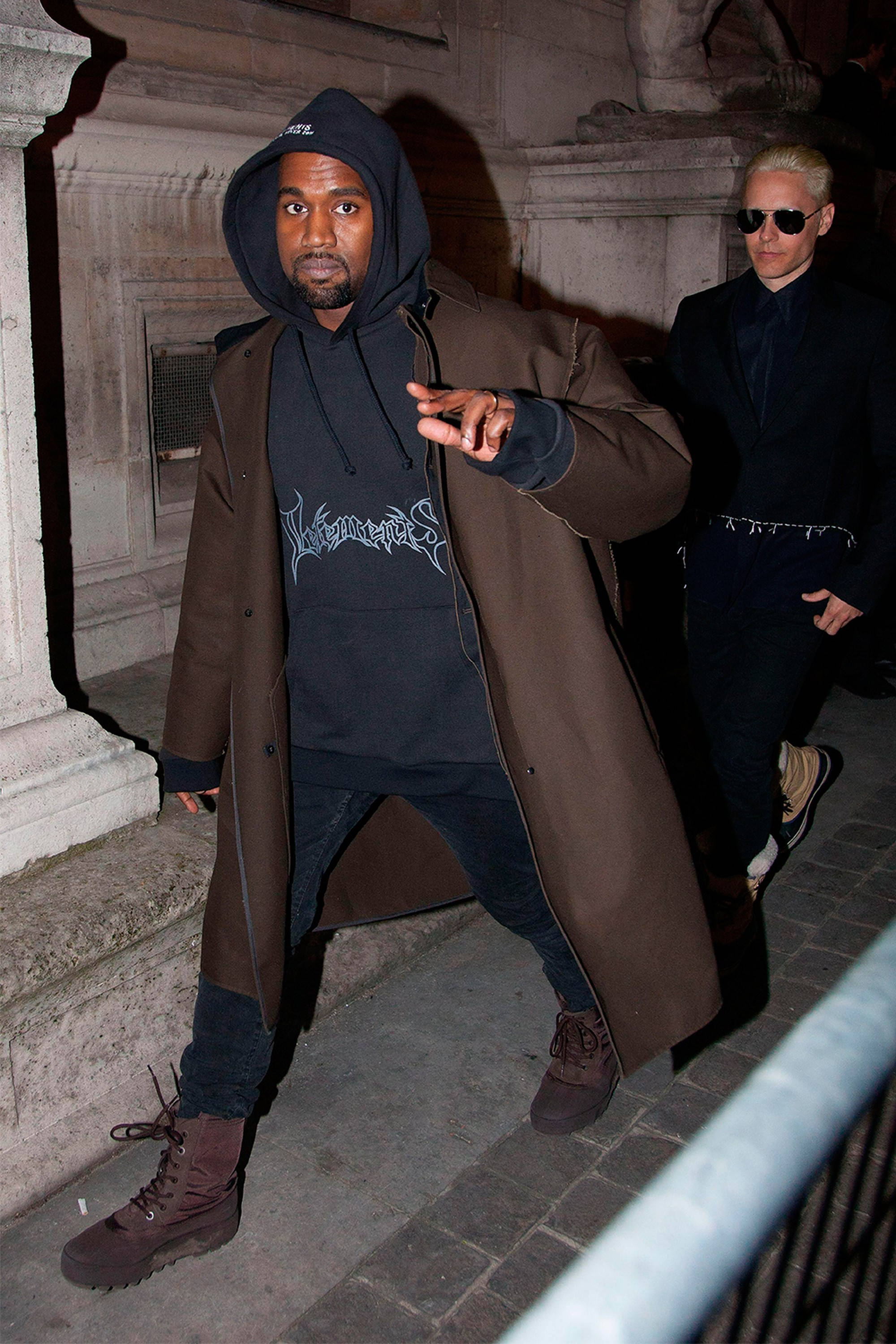 Kanye West 身穿 Vetements 連帽衫 壁紙 — Kanye West 穿著棕色外套和連帽衫，在夜間自信地走在石鋪的城市街道上，身後有一位同伴。