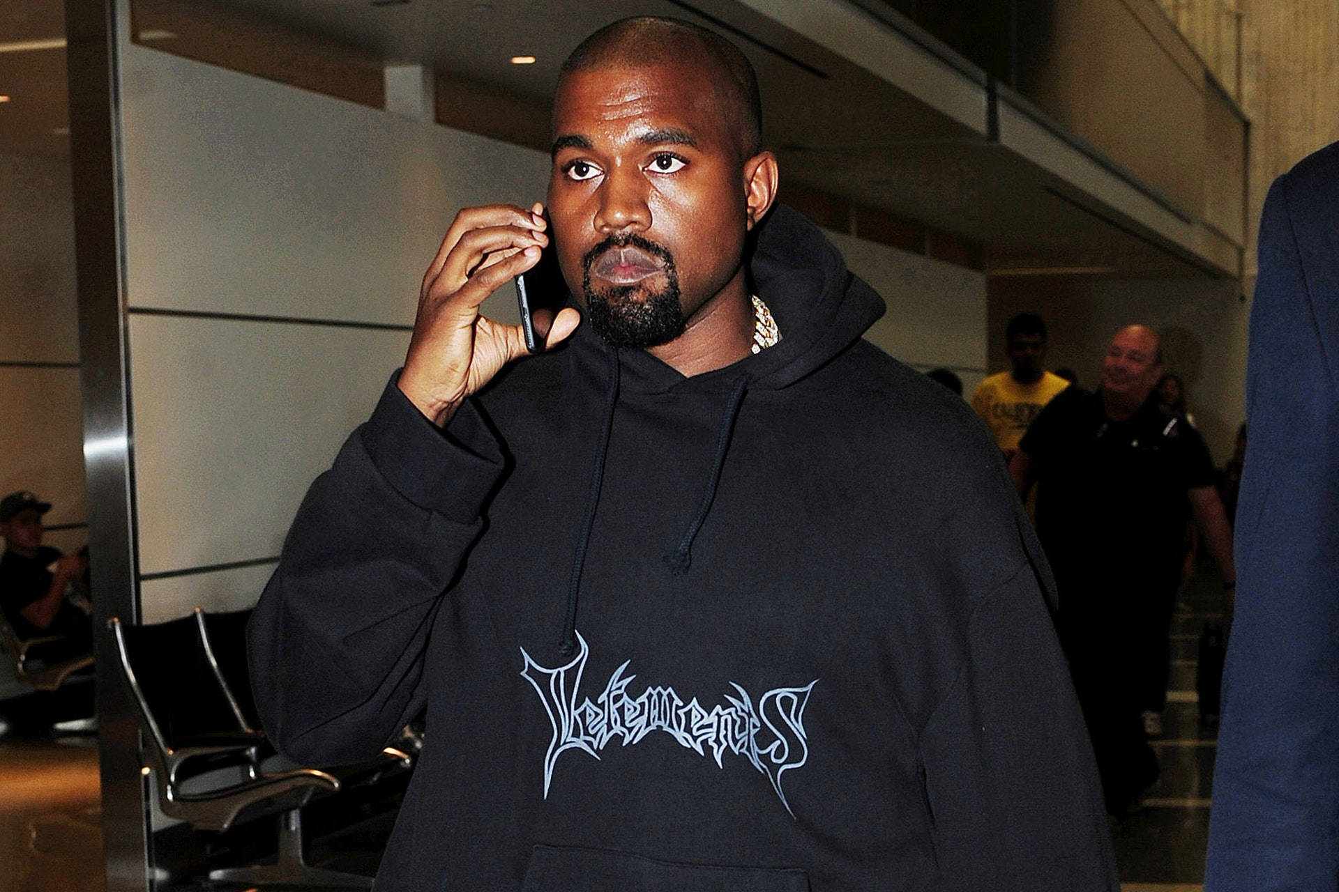 Vetements Kanye West 壁紙 — Kanye West 身穿黑色連帽衛衣，上面有白色塗鴉風格文字，在現代室內走廊講電話。