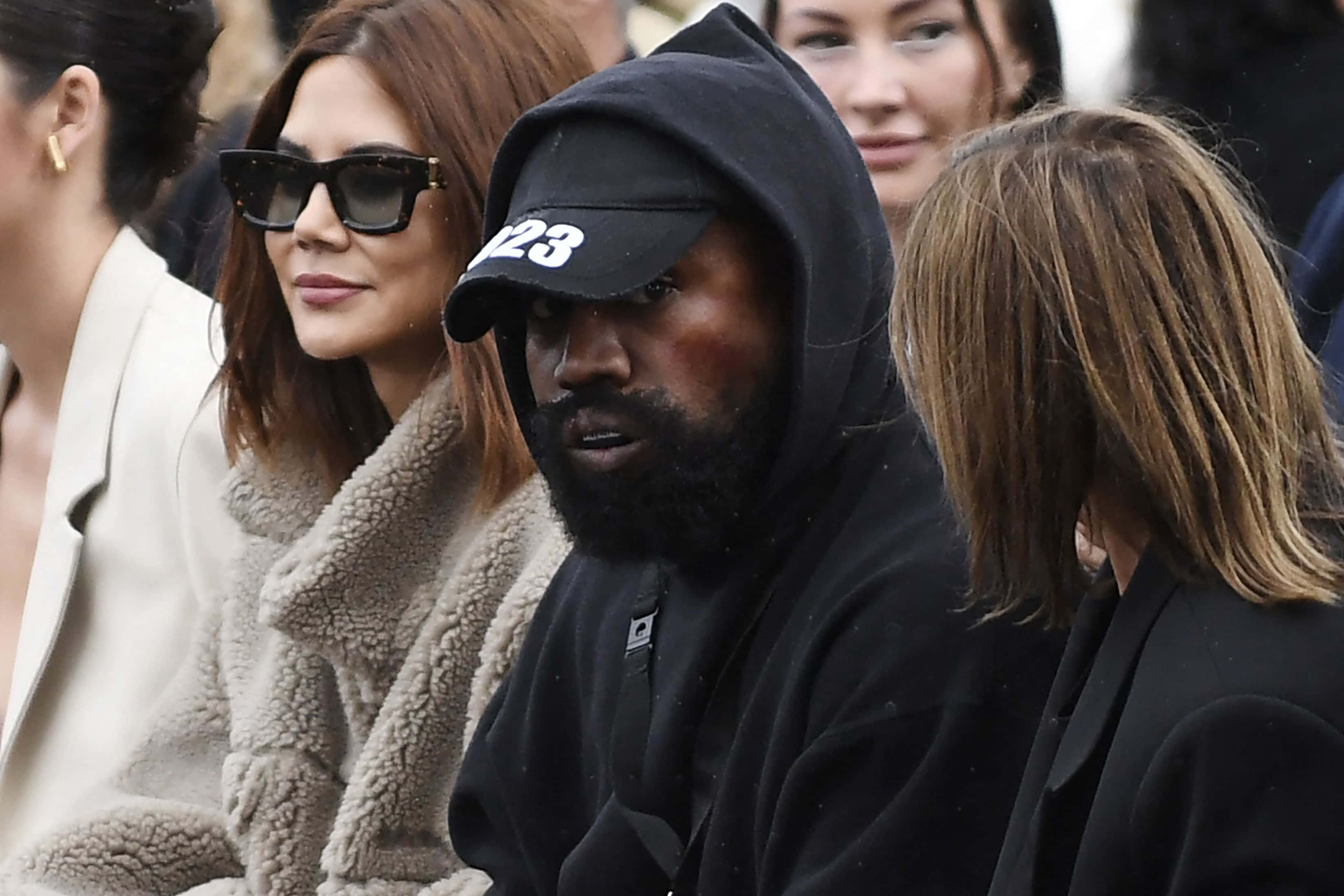 Kanye 桌布 壁紙 — Kanye West 身穿黑色連帽衫和帽子，坐在身穿米色羊羔毛外套的 Kim Kardashian 旁邊，出席公開活動。