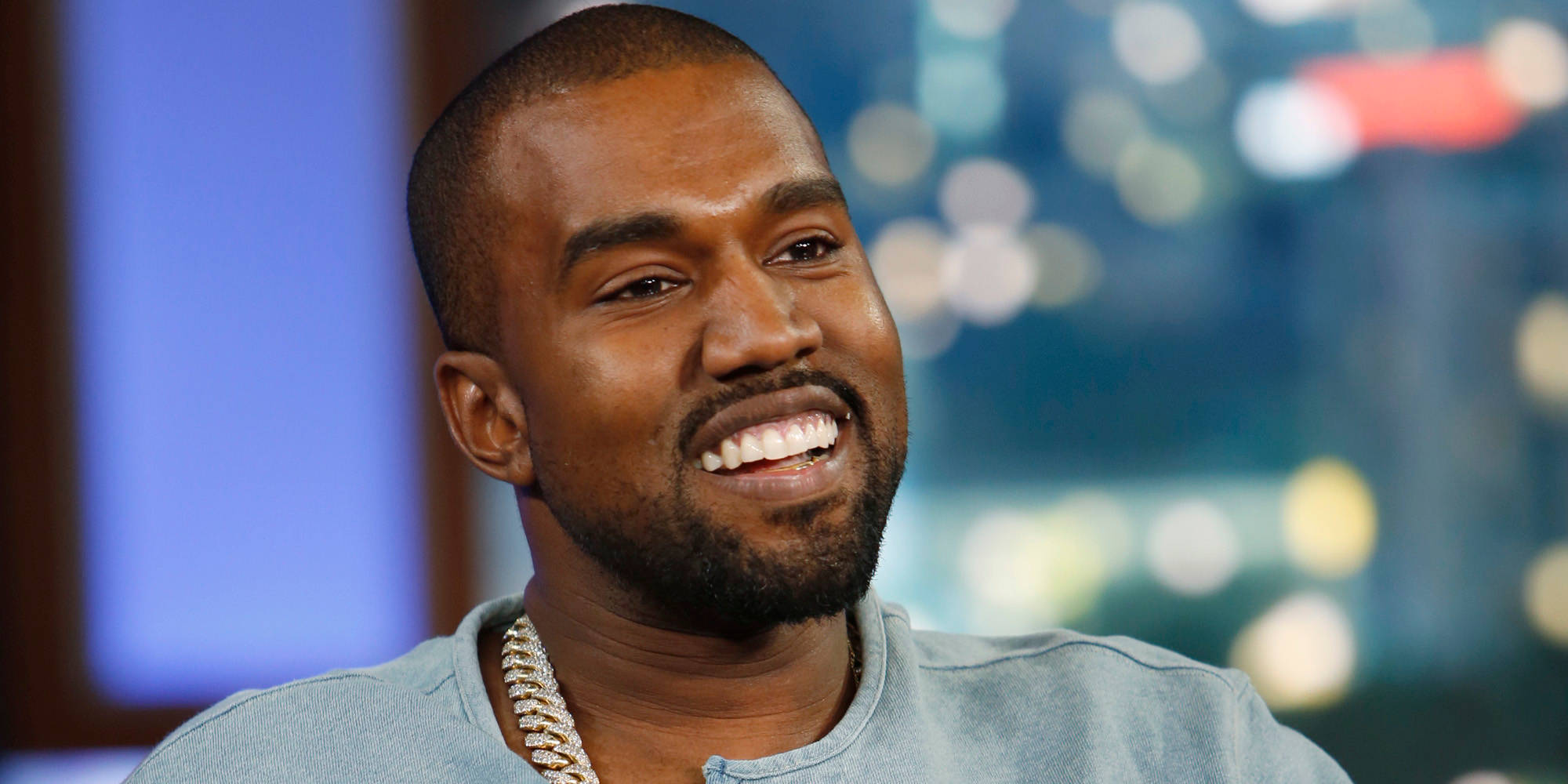 Kanye West微笑 壁紙 — Kanye West 戴著鏈條項鍊，在模糊的城市景觀背景下燦爛地微笑。