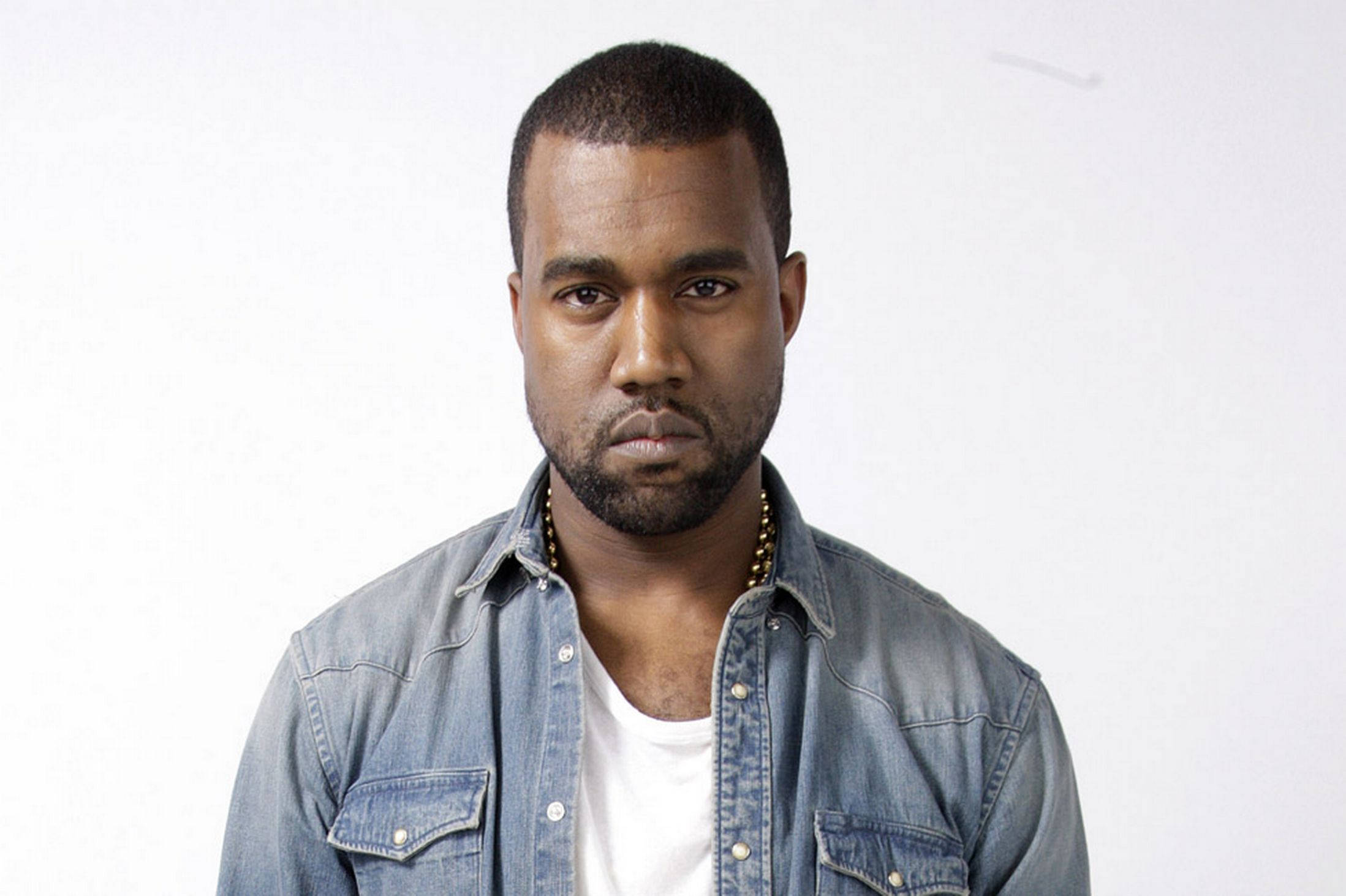 Kanye West極簡攝影 壁紙 — 一個表情嚴肅的男人，身穿牛仔襯衫內搭白色T恤，背景為純白色。
