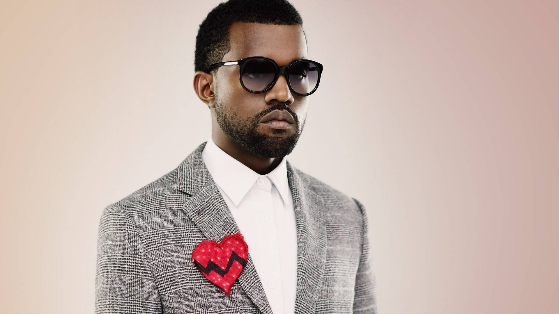 Kanye West穿灰色西裝 壁紙 — 一位時尚男士身穿灰色西裝和太陽眼鏡，在柔和的粉紅色背景上佩戴紅色破碎心形別針。