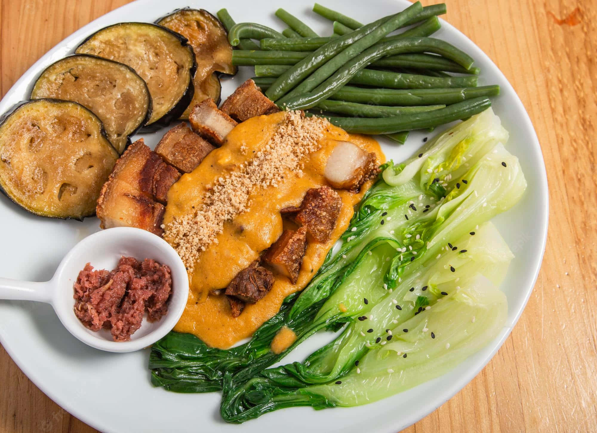 Kare-Kare 的新視角 壁紙 — 一個充滿活力的烤茄子、青豆和脆皮豬肉配奶油醬和芝麻的菜餚，放在木桌上。