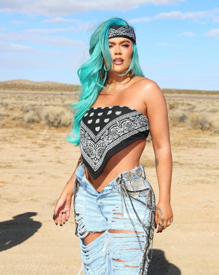 Karol G《Location》歌曲海報 壁紙 — 一位擁有充滿活力的綠松石色頭髮和頭巾上衣的女性，自信地站在陽光明媚的沙漠景觀中。
